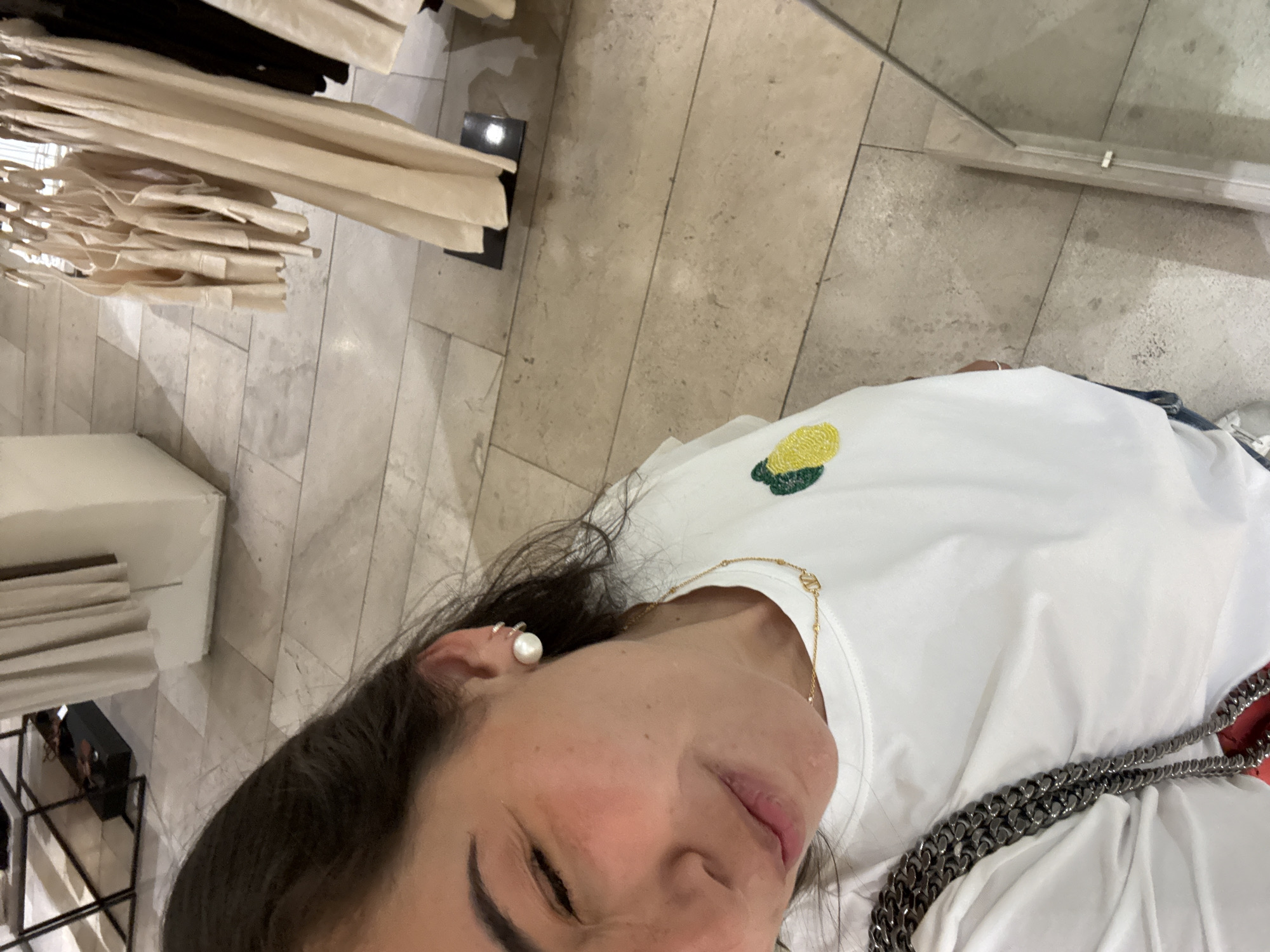 The image shows a woman wearing a white t-shirt with a corn embroidery, pearl earring and a silver chain bag strap. She appears to be in a store in Puebla, Mexico, as suggested by the mall-like environment with clothing racks and marble flooring.