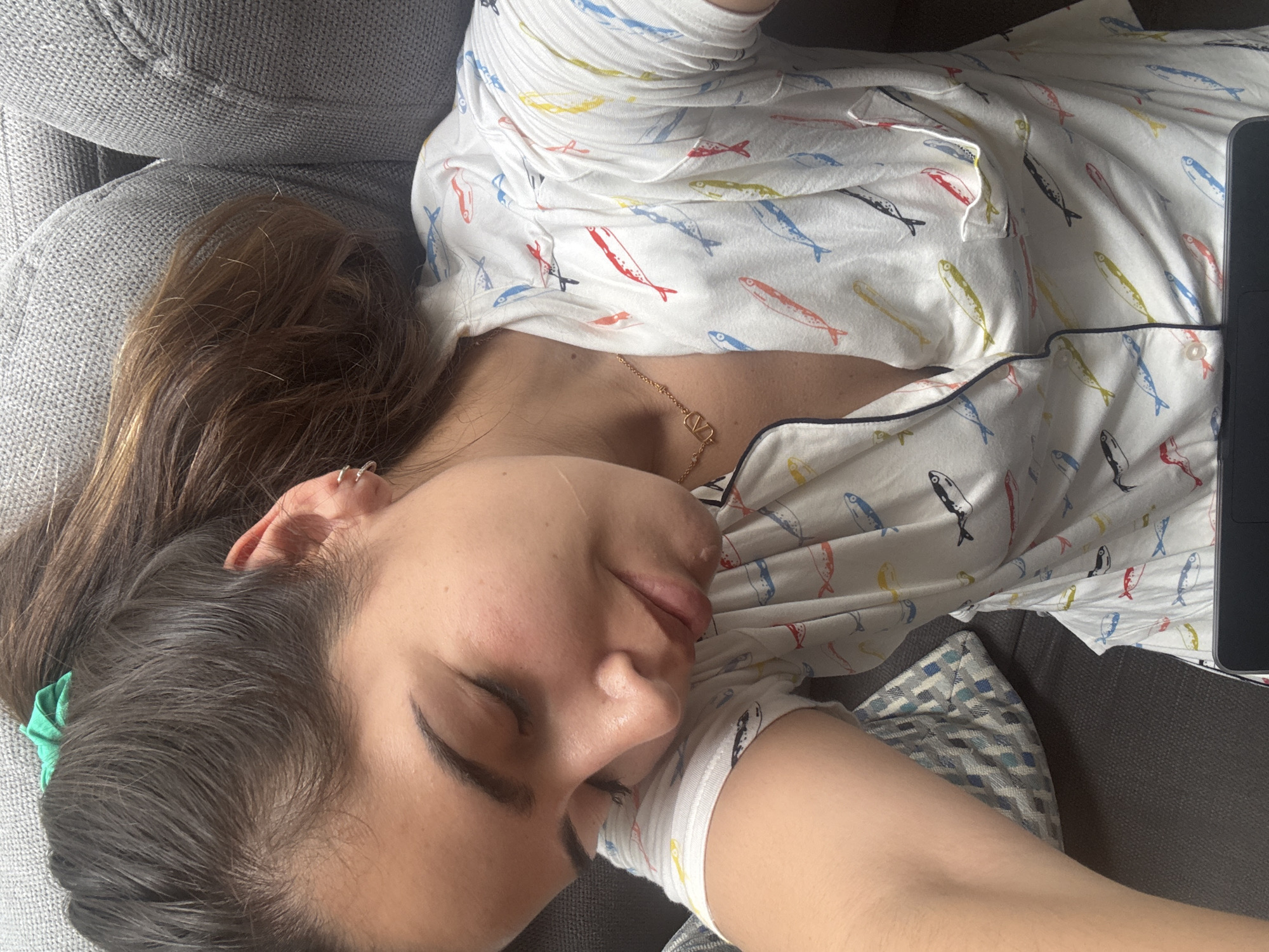 A woman with brown hair and fair skin is lying on a gray couch. She is wearing pajamas with a fish pattern and a gold necklace. A laptop is visible to her right. Her eyes are closed, suggesting she is sleeping or resting. A green hair tie is in her hair. No details indicate a location in San Bernardino Tlaxcalancingo, Mexico.