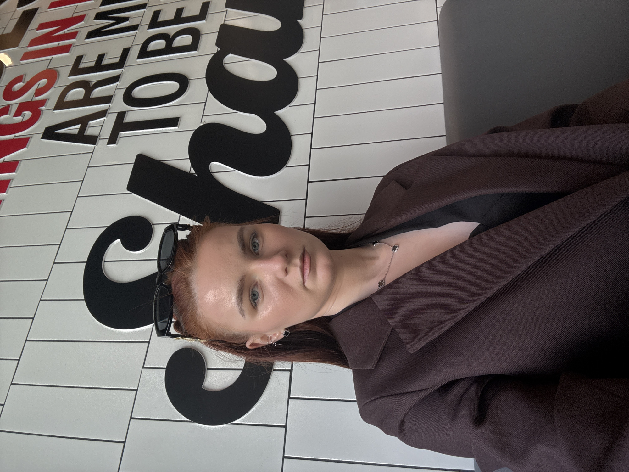 A woman is seated in front of a wall with the word "SCHOOL" written on it in large, black lettering against a white tiled background. The lettering is incomplete, obscured by her presence. The phrase "THINGS ARE TO BE" is visible above "SCHOOL" in red and black.
She is wearing a brown blazer, a necklace and has sunglasses perched atop her head. She is looking directly at the camera.
Based on the available information, no definitive information supports the claim that the location is in the city of Novoe Devyatkino, Russia.