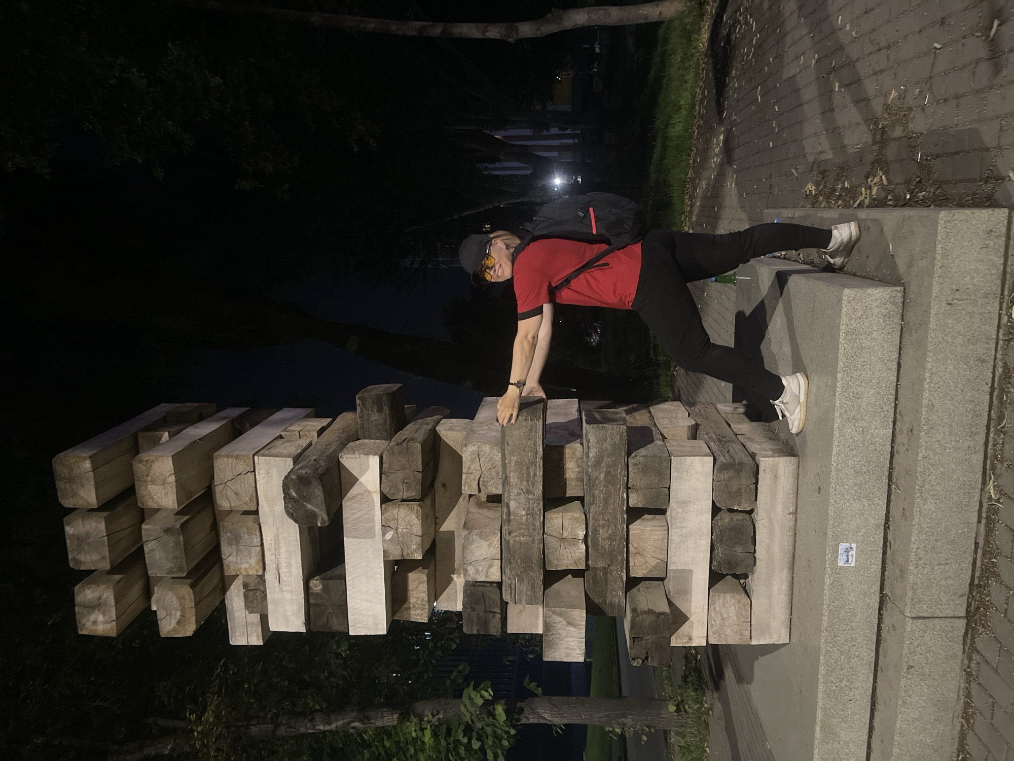 A person wearing a red t-shirt, black pants, a black baseball cap, and sunglasses is standing next to a large Jenga-like sculpture made of wooden beams. The sculpture is tall and narrow, with beams stacked unevenly, resembling a giant game of Jenga. The person is leaning against the sculpture with their hands on it and their body angled towards the camera. They are also wearing a black backpack. The scene is outdoors at night, with trees and dark shadows in the background. A paved walkway is visible in the foreground. There is a small sticker on the base of the sculpture. The location is identified as Novokuybyshevsk, Russia.