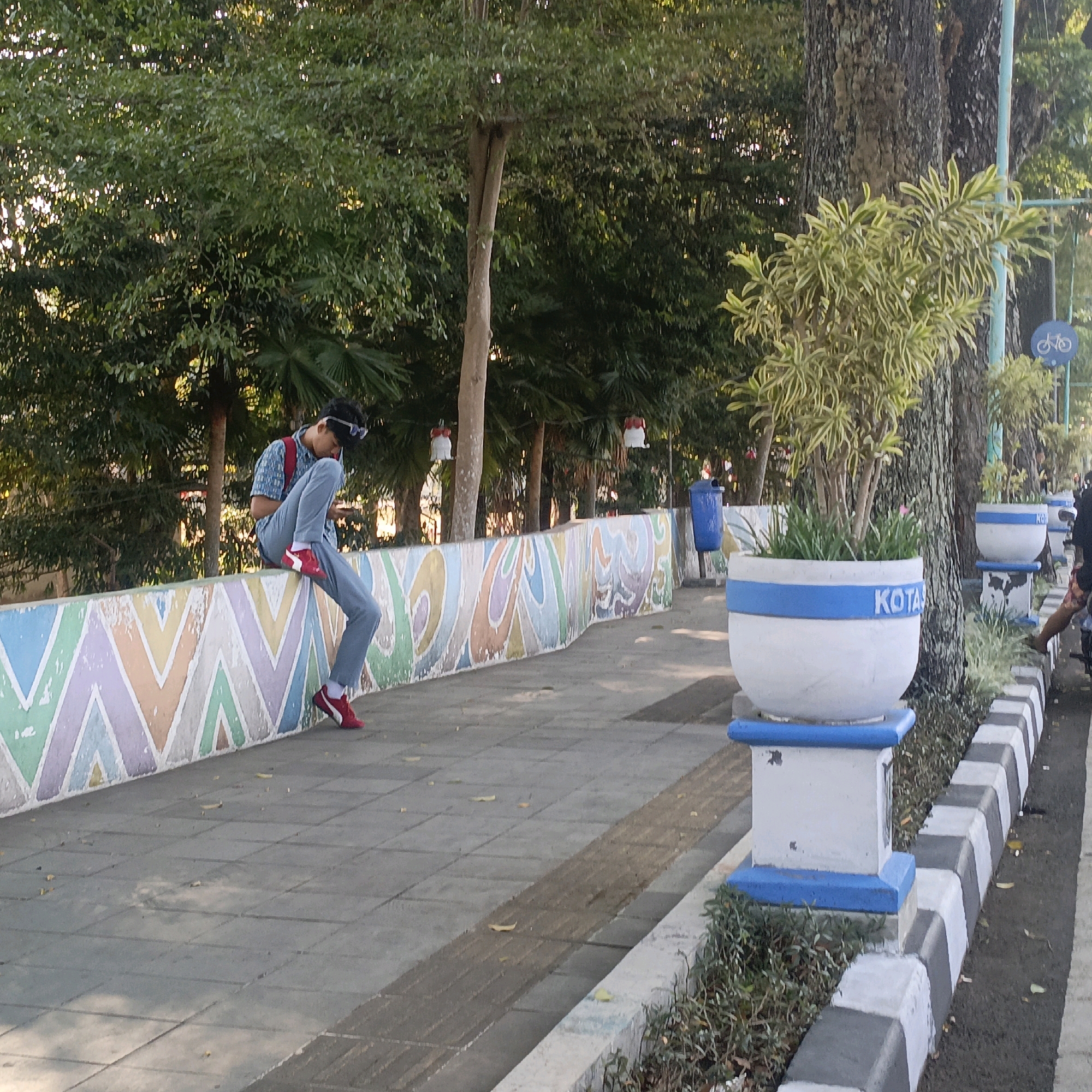 This image captures a sunny outdoor scene in Salatiga, Indonesia. A young man is seated on a low, decorative wall, leaning forward and looking at his phone. He is wearing a blue patterned shirt, grey pants, and red sneakers. The wall he is sitting on is decorated with a repeating pattern of colorful, zig-zagging lines in shades of white, purple, green, and orange. Behind the wall, a paved walkway is visible, with a tactile paving strip for the visually impaired running alongside it. Beyond the walkway, several large trees with dense green foliage provide shade. In the foreground, a white planter with a blue rim, containing green foliage, sits on a blue and white base. Further down the walkway, another similar planter is visible, and to the right, a black and white striped curb marks the edge of the road. A blue trash bin is also present near the wall. The overall environment suggests a public park or a pedestrian area in the city.