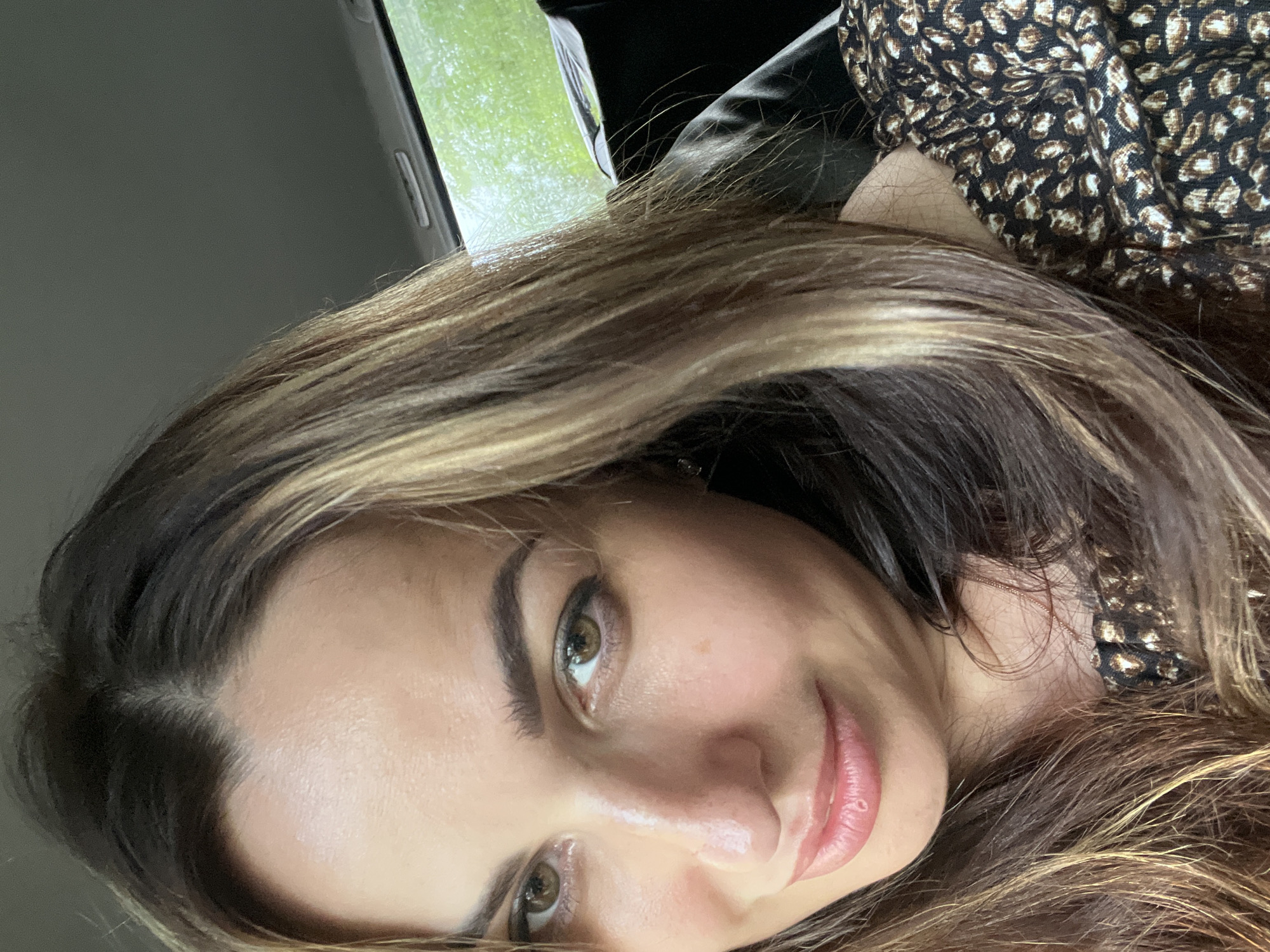 A close-up, waist-up, candid shot captures a woman with long, layered brown hair with blonde highlights. She is indoors, likely in a car, as indicated by the visible car ceiling and window with greenery outside. The woman has fair skin, brown eyes, and is wearing a black top with a brown leopard print. She is looking towards the camera and smiling slightly. The lighting suggests it is daytime.