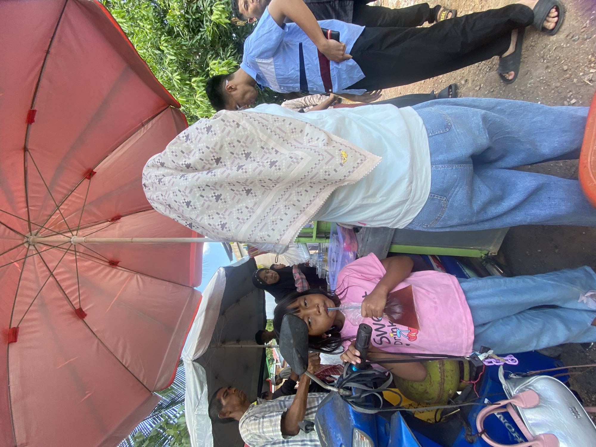 A young girl in a pink t-shirt and blue pants drinks from a plastic bag of brown liquid with a straw. She is standing in front of a blue motorcycle, under a large pink umbrella. Behind her, a woman in a headscarf and a man in a light blue shirt are partially visible. A man in a plaid shirt stands to the left, looking forward. The scene appears to be an outdoor market or street food stall, likely in Banjarbaru, Indonesia, given the context. The ground is dirt, and trees are visible in the background. A silver handbag with pink straps rests on the ground near the motorcycle.