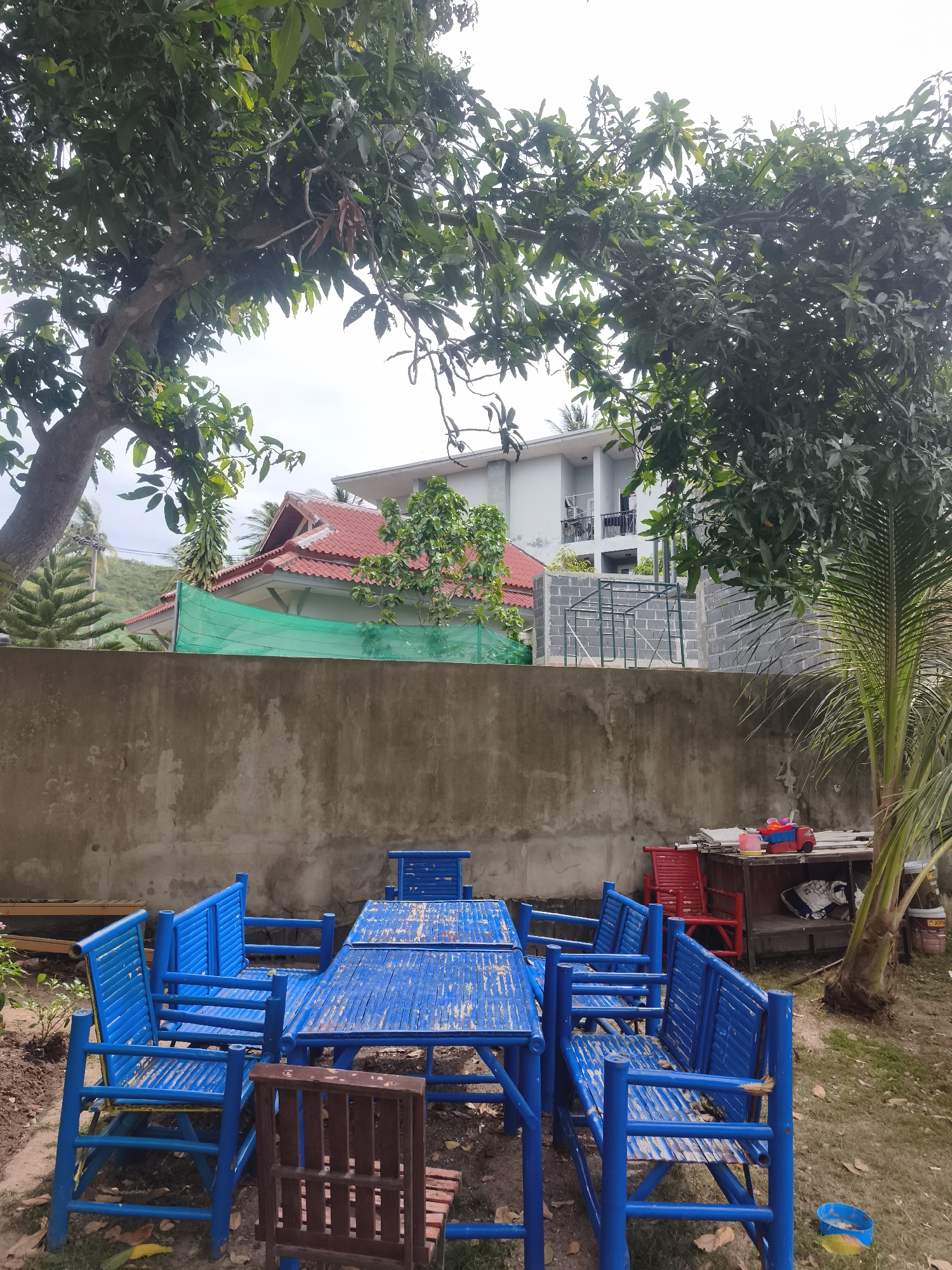 In the city of Ko Samui District, Thailand, a medium shot captures an outdoor seating area. Dominating the foreground are several blue bamboo tables and benches, showing signs of wear. A dark brown wooden chair is positioned in front of one of the tables. Behind the seating area, a textured, gray concrete wall forms a barrier. Above the wall, the upper stories of a light-colored building with balconies are visible, with a red-tiled roof to the left. Lush green foliage from large trees arches over the scene, with a palm tree on the right. The environment appears to be a garden or yard, with scattered leaves on the ground and some construction materials to the right. The sky is overcast.