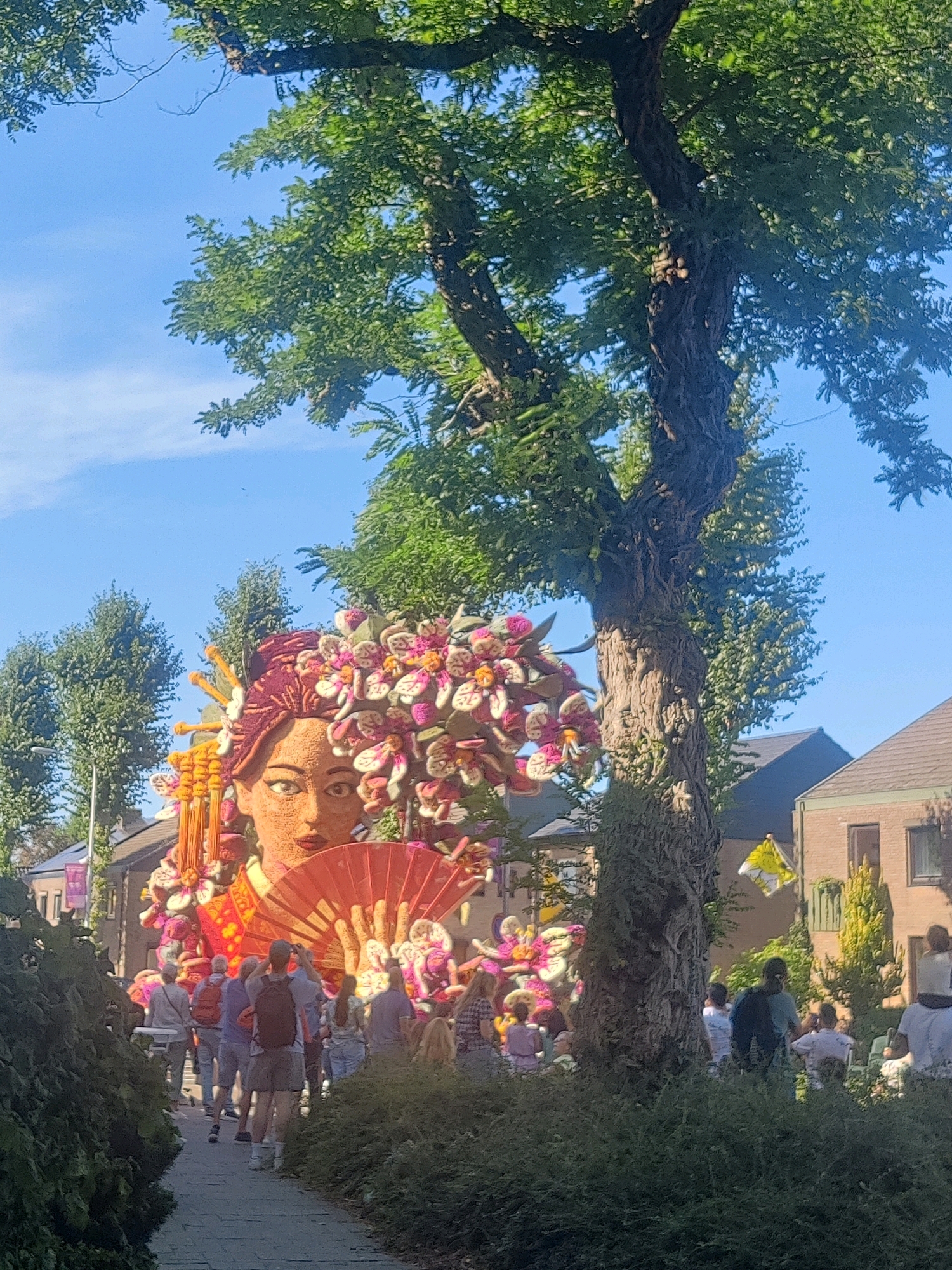 The image depicts a flower parade float resembling a woman's face with an elaborate floral headdress and a large orange fan held in her hand. The float is decorated with various flowers, predominantly in shades of pink, white, and red. A crowd of people is gathered on a paved pathway, observing the float. Many individuals are holding cameras or phones, indicating they are documenting the event. The parade appears to be taking place in a residential area, with houses visible in the background. Tall green trees with lush foliage frame the scene, and the sky is a clear blue. The overall atmosphere suggests a festive outdoor event, likely part of the annual Zundert Flower Parade, a tradition held in the city of Zundert, Netherlands, known for its impressive floral floats.