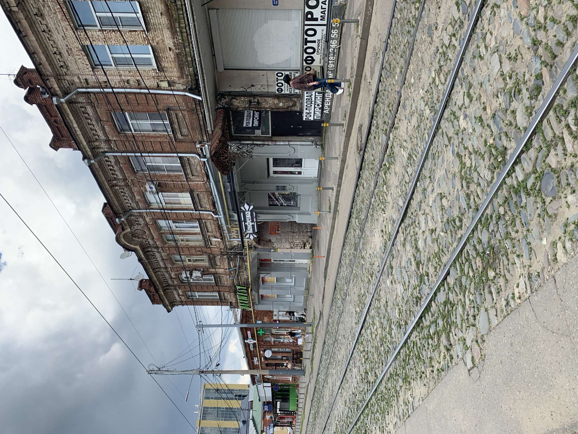 Here is a description of the image:
The image shows a street scene with tram tracks running along a cobblestone surface. A multi-story brick building stands to the left, featuring a mix of windows and architectural details. Several signs are visible on the building's facade, including one for a "PHOTO" shop and another for "ROCK-PODVAL" with the word "PIRSING" (piercing) underneath. A person is standing near the "PHOTO" shop sign. Other signs in Russian, including one for "Konfetka" (Candy), can be seen down the street. Power lines run overhead, and a cloudy sky forms the backdrop. Further down the street, glimpses of modern buildings are visible.