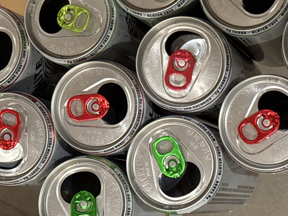 The image shows a collection of open beverage cans viewed from above. Some cans have red tabs, while others have green tabs. Text is visible on the cans, including phrases like "Gluten Free," "Made Pure," "100 Calories," "2g Carbs," and "MAME NY VTSO HUA." The cans are arranged close together, creating a pattern of silver, red, and green.
