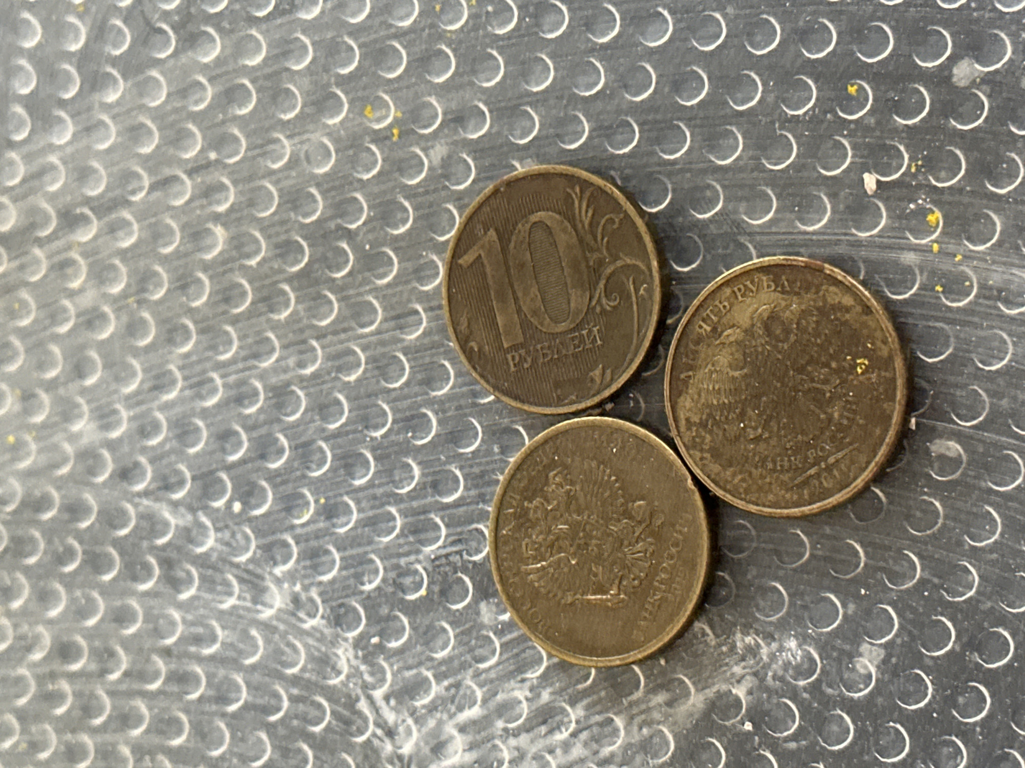 Three Russian coins are arranged in a triangular formation on a dark, textured surface with a repeating pattern of small, raised, crescent-shaped bumps. The coin on the left is partially visible, showing the double-headed eagle emblem and text that appears to include "БАНК РОССИИ" and the year "2019". The coin at the top right clearly displays the numeral "10" and the word "РУБЛЕЙ" (rubles) in Cyrillic script. The coin at the bottom right also features Cyrillic text and an emblem, though it is less distinct. The surface appears to be some form of metallic or plastic grating. There are no people or any discernible elements that would place this scene within the city of Nevinnomyssk, Russia. The lighting suggests an overhead light source, creating subtle shadows and highlights on the coins and the textured surface.