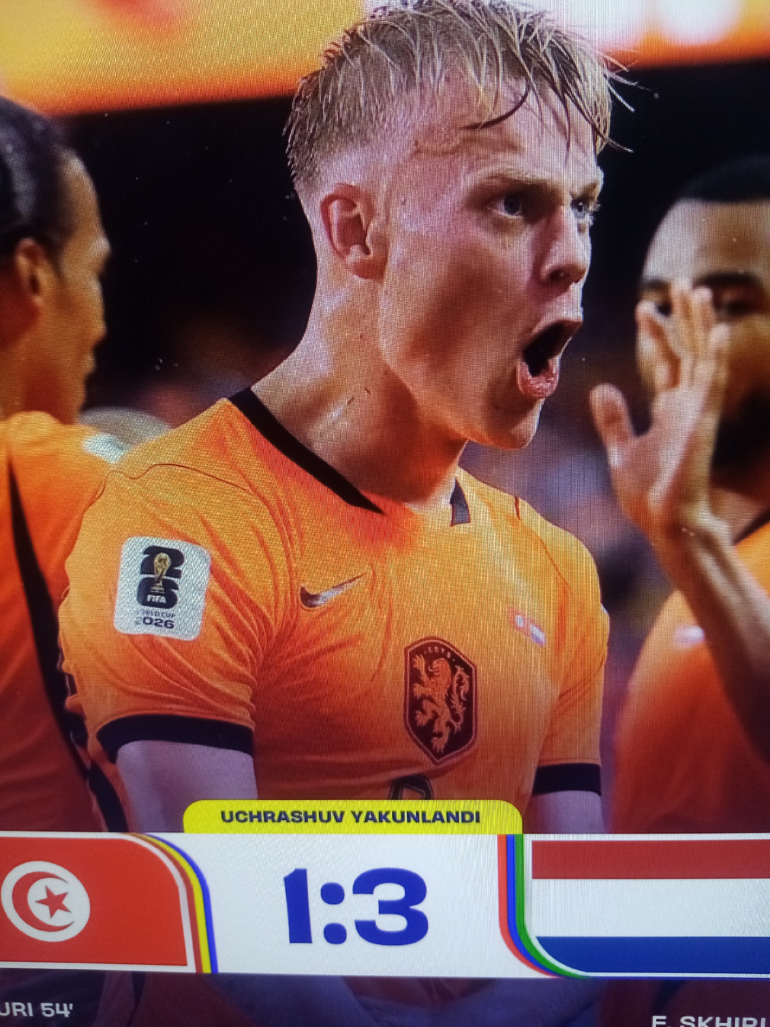 The image captures a close-up of a young male athlete, likely a soccer player, with blond hair styled in a short fade. He is wearing an orange jersey with a crest and a Nike logo. His mouth is open in a yell or cheer, conveying strong emotion. Behind him, two other individuals are partially visible, also appearing to be athletes, one with braided hair and the other in profile.
A graphic overlay at the bottom of the image displays a soccer match score of "1:3". To the left of the score is a flag with a red background and a white crescent moon and star, representing Tunisia. To the right of the score is the flag of the Netherlands, featuring red, white, and blue horizontal stripes. The text "UCHRASHUV YAKUNLANDI" is visible above the score, which translates to "Match Ended" in Uzbek, indicating the event is a soccer match. The presence of the Uzbekistan text suggests the context is within Chirakchi, Uzbekistan. The lighting appears to be artificial, typical of a stadium or broadcast setting. The overall scene suggests a moment of intense emotion following the conclusion of a soccer game, with the score indicating a Dutch victory over Tunisia.