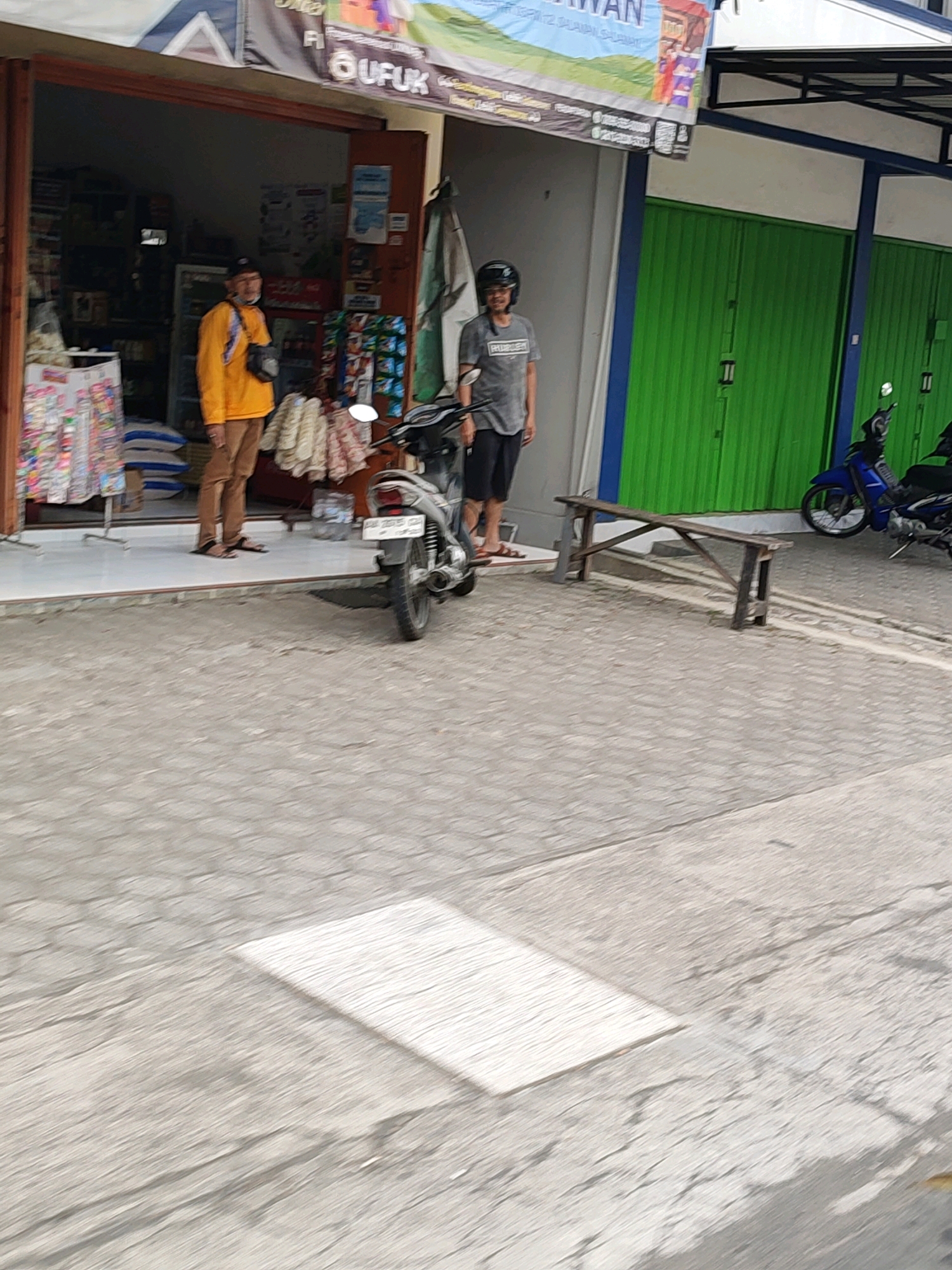 In the city of Salaman, Indonesia, two men stand outside a shop. One man, wearing a yellow long-sleeved shirt and brown pants, stands in the doorway of the shop, which is filled with shelves of merchandise. The other man, wearing a gray t-shirt and black shorts, stands on the sidewalk to the right of the doorway, near a motorcycle. A blue motorcycle is parked to the right of the second man, and a wooden bench sits on the sidewalk between them. The ground is paved with grey cobblestones, and the shop has a blue awning. A banner is visible above the shop, with text that is partially obscured.