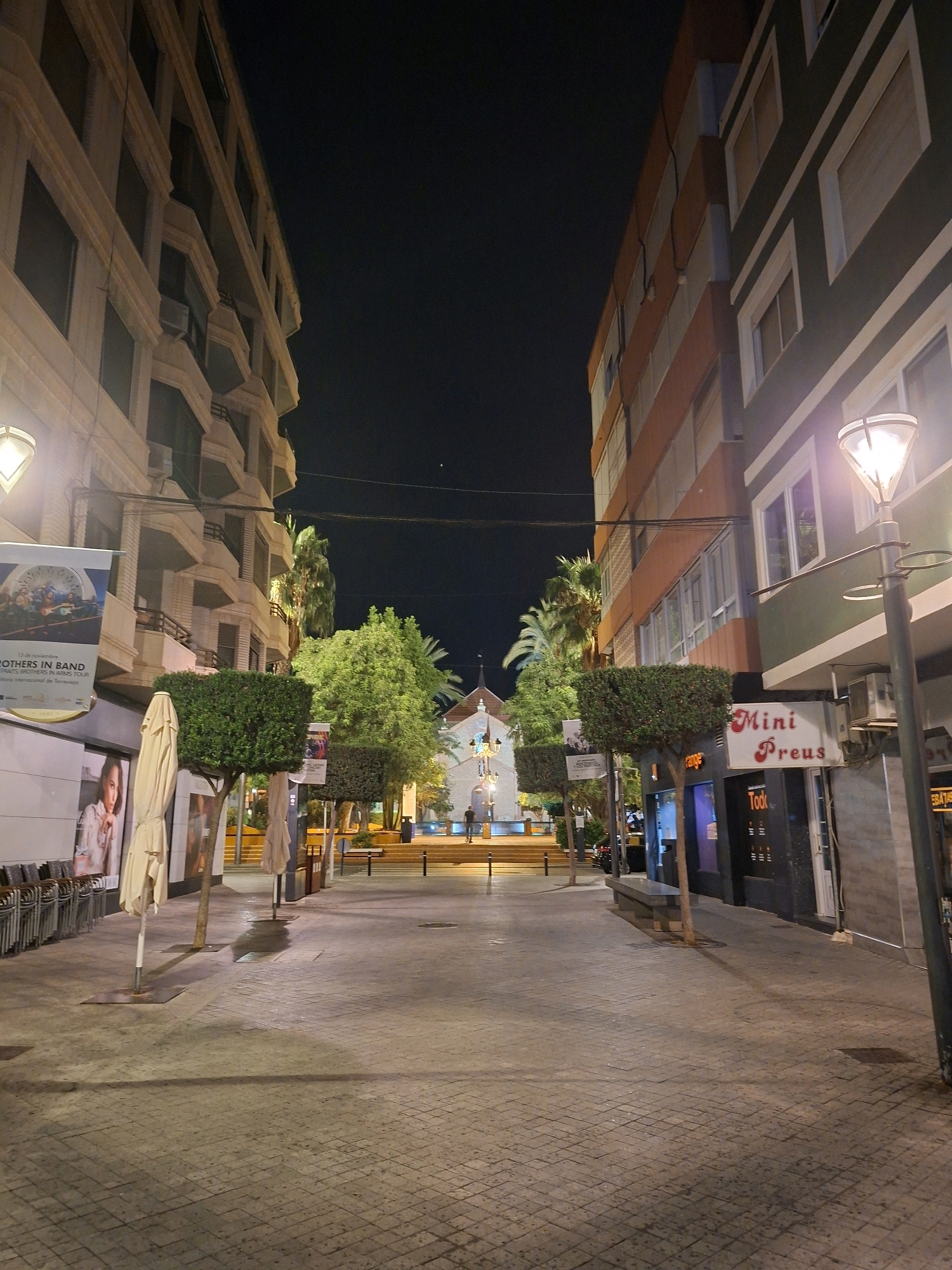 This is a nighttime, eye-level, medium-shot photograph of a pedestrian street in Torrevieja, Spain. The street is lined with multi-story buildings on both sides, creating a canyon effect. The building on the left is a light-colored building with balconies, while the building on the right is a darker building with orange and green sections.
In the foreground, the street is paved with patterned cobblestones. There are several trees and planters along the sides of the street. On the left, a stack of chairs and tables are visible outside a building, and a large poster hangs on the wall. On the right, a street lamp illuminates the scene, and a sign reads "Mini Preus". Further down the street, a small white building with a steeple is visible, possibly a church.
There are a few people walking in the distance, but no significant interactions or actions are clearly depicted. The overall atmosphere is calm and quiet, typical of a street at night. The lighting suggests artificial illumination from street lamps and building lights.