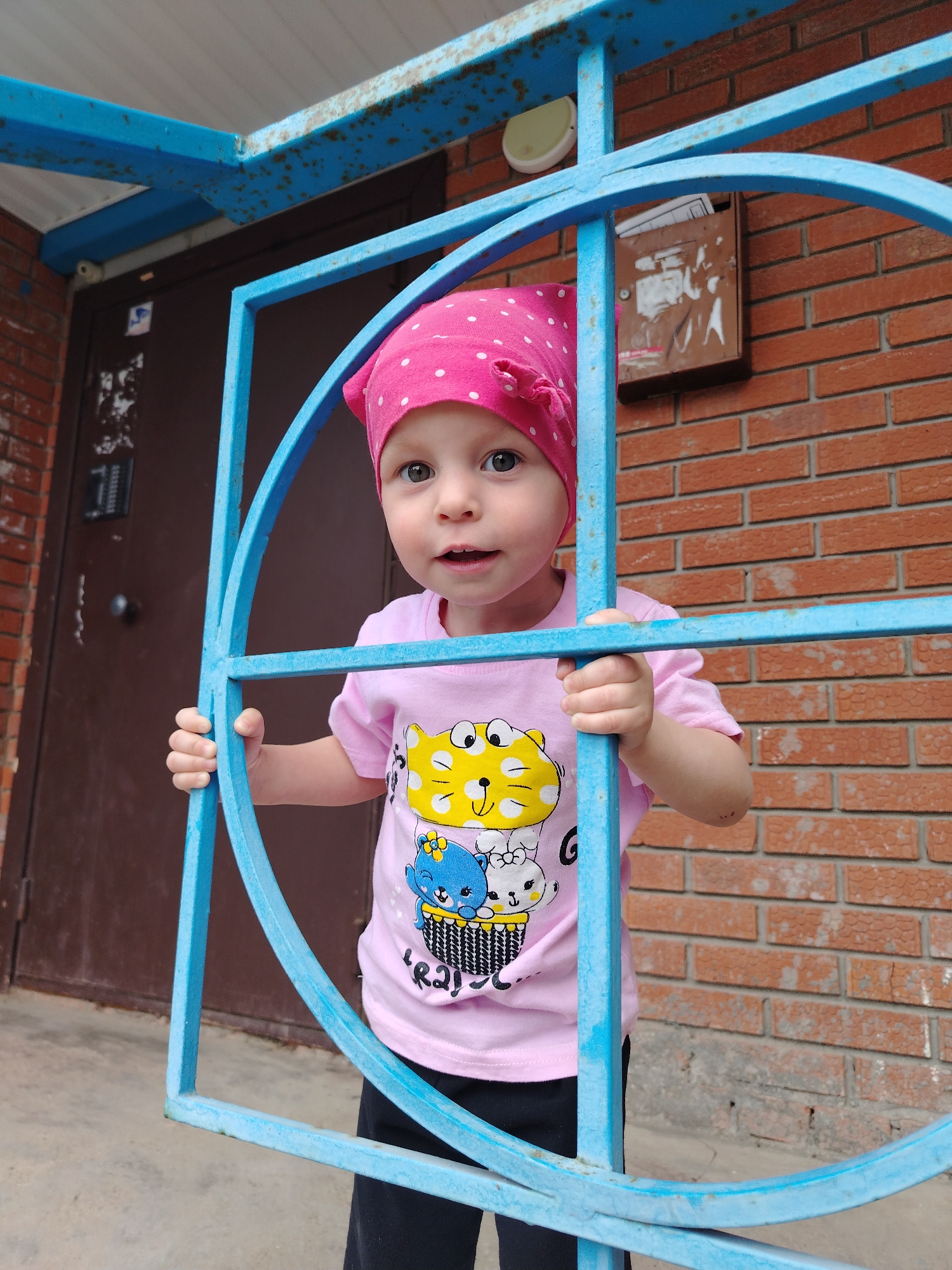 A young child wearing a pink polka-dot headscarf and a pink T-shirt with cartoon animal images is standing behind a blue metal structure resembling a gate or barrier. The child is looking directly at the camera with an expression of curiosity or surprise. The setting appears to be outdoors in front of a brick building with a brown metal door and a mailbox. The metal structure shows signs of rust or weathering. The location is in the city of Chaikovsky, Russia.