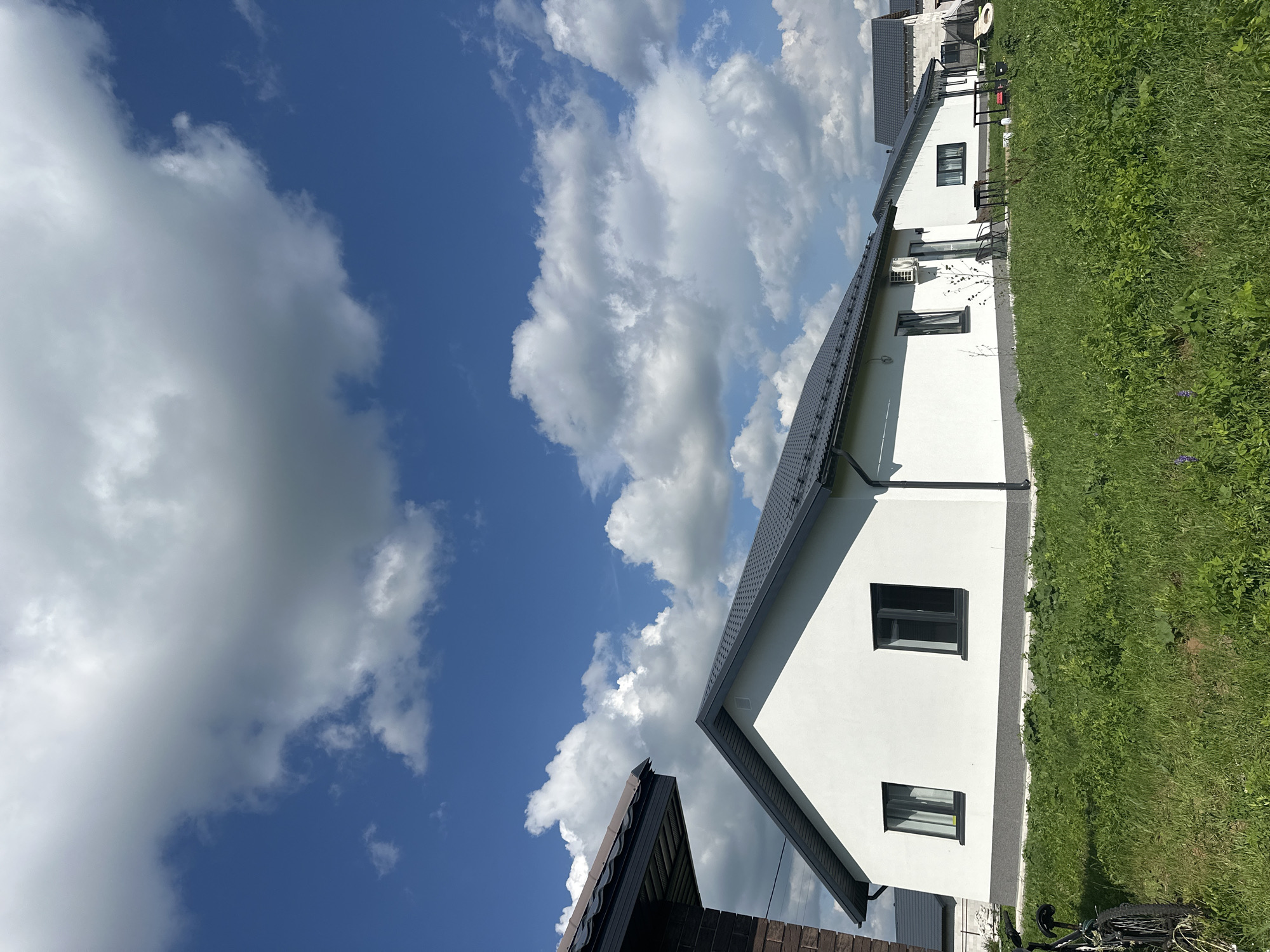 The image depicts a bright, sunny day in Mokino, Russia, with a clear blue sky dotted with fluffy white clouds. In the foreground, a lush green lawn stretches out, with a bicycle parked on the left side. Several modern, white houses with dark gray roofs are visible, some under construction. The houses are single-story structures with clean lines and dark-framed windows. One house has an air conditioning unit mounted on its exterior wall. In the background, more houses and some building materials, including what appears to be a trampoline, can be seen. The overall impression is one of a developing residential area on a beautiful summer day.