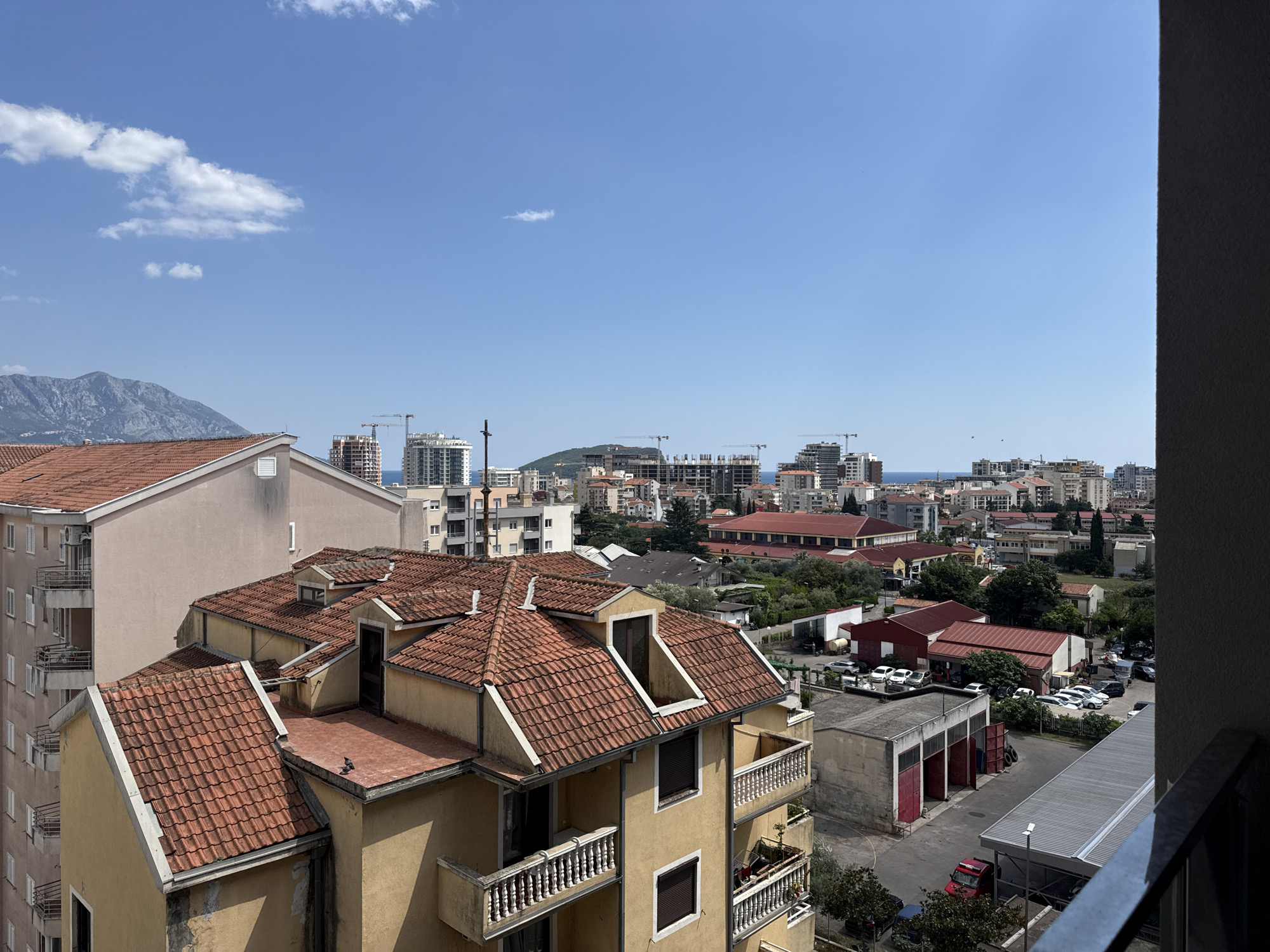 The image displays a cityscape of Budva, Montenegro, under a clear blue sky with a few scattered clouds. In the foreground, a multi-story building with a reddish-brown tiled roof and beige exterior is prominent. This building features balconies with white railings and dark-shuttered windows. Adjacent to it, a taller, pale pink building with similar balconies and white railings is visible.
Beyond the foreground buildings, the city unfolds with a mix of architectural styles. Several mid-rise buildings and apartment complexes are present, with some appearing to be under construction, indicated by visible cranes. A cluster of red-roofed structures, possibly commercial or industrial buildings, can be seen, along with a parking lot filled with various vehicles, including cars and a red truck. In the distance, a mountainous landscape forms the backdrop, meeting the horizon where the sea is faintly visible. The overall scene is one of urban development against a natural, mountainous coastline.