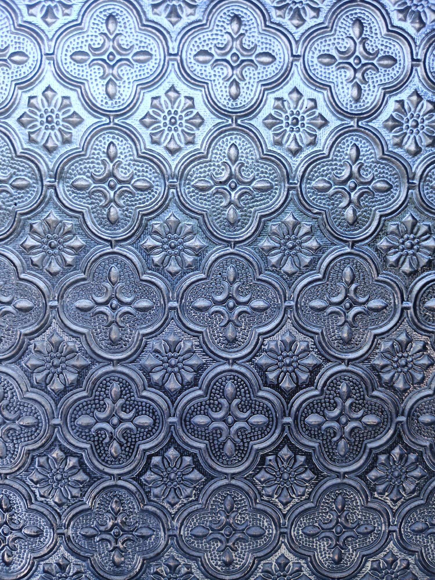 The image is a close-up, full-frame shot of a decorative patterned glass, possibly stained glass, with a blue-grey hue. The pattern features repeating floral and geometric motifs within a quatrefoil-like structure. The glass is textured with raised and recessed areas, creating a relief effect. There are no people or specific environmental details visible, indicating it is a detail shot of an architectural element. The pattern is complex and symmetrical. Without additional context, the location in "Unknown, Indonesia" is an external label and not derivable from the visual information.