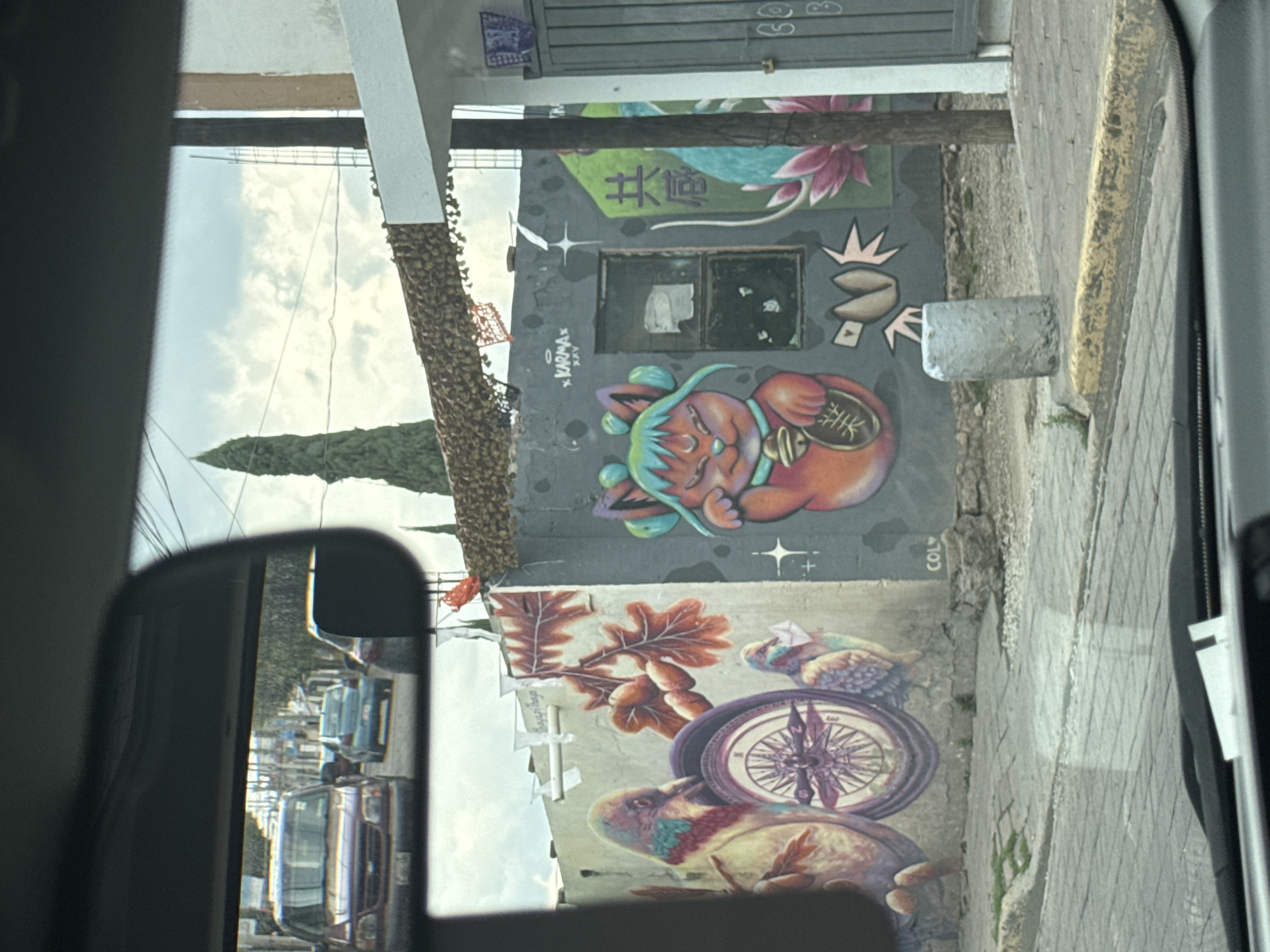 The image shows a street scene in San Andrés Cholula, Mexico, viewed from inside a vehicle. A building wall is visible, adorned with colorful street art. The artwork features elements such as a stylized cat-like figure with a coin, flowers, leaves, a compass, and birds. The wall also has graffiti tags and markings. The sidewalk and street are visible, with a curb separating them. In the vehicle's side mirror, a row of parked vehicles can be seen along the street. The sky is visible above the buildings.
