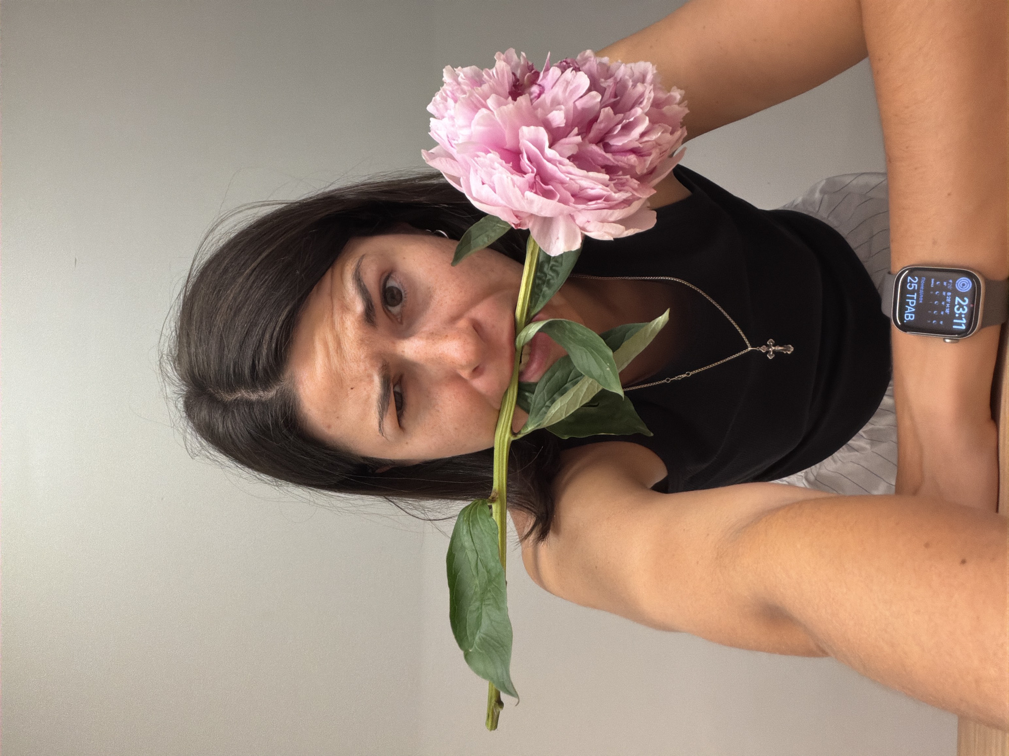 A woman is holding a pink peony in her mouth and winking at the camera. She is wearing a black top and a silver cross necklace. An Apple Watch is visible on her left wrist, displaying the time 23:11 and the date 25th of some month. The background is a plain, light gray wall. There is no indication of the location being Budva, Montenegro, or any specific city.