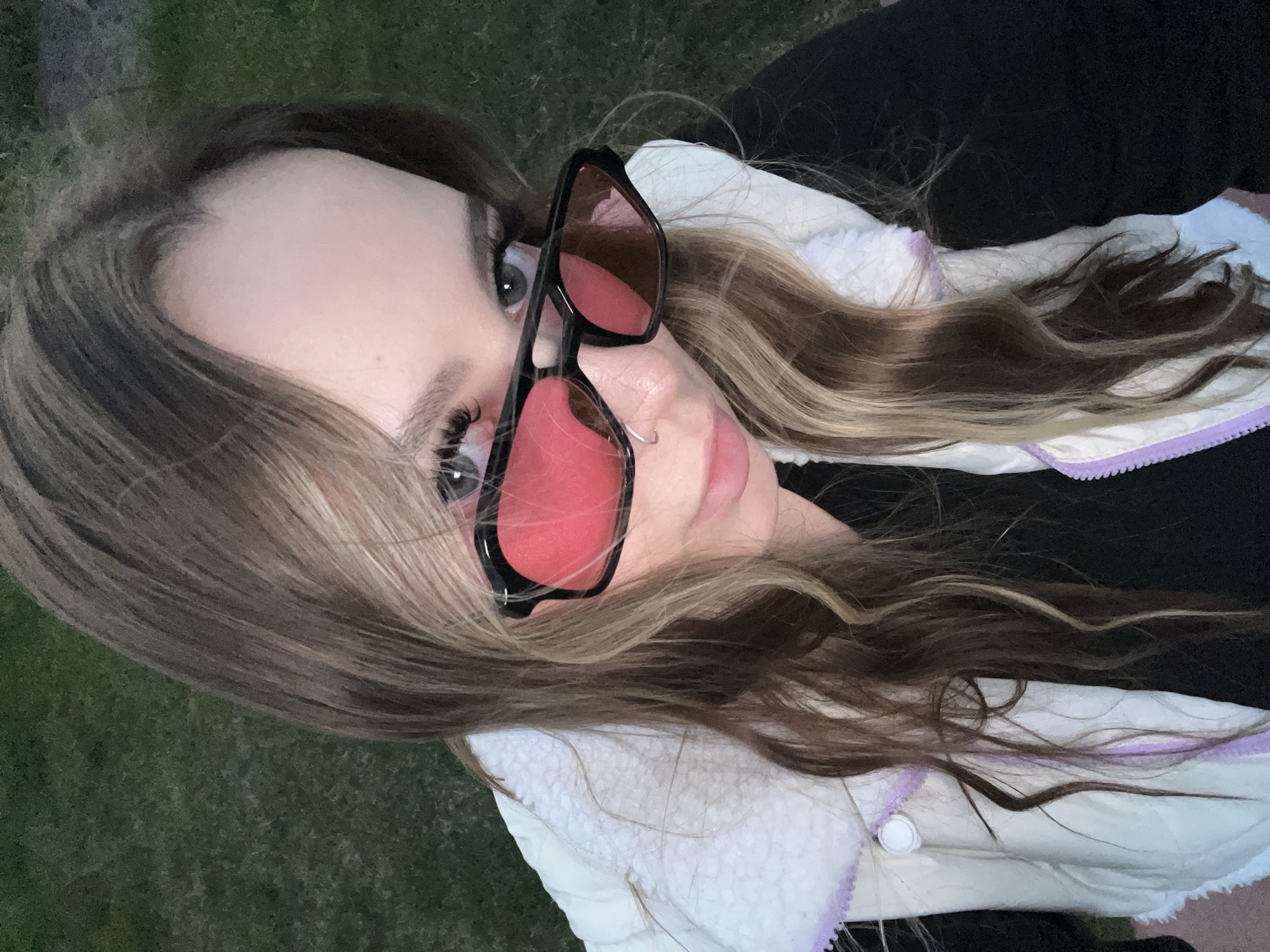 A woman with long, brown and blonde hair is lying on the grass, wearing sunglasses with red lenses and a white jacket with a black shirt underneath. The location cannot be determined as Krasnodar, Russia.