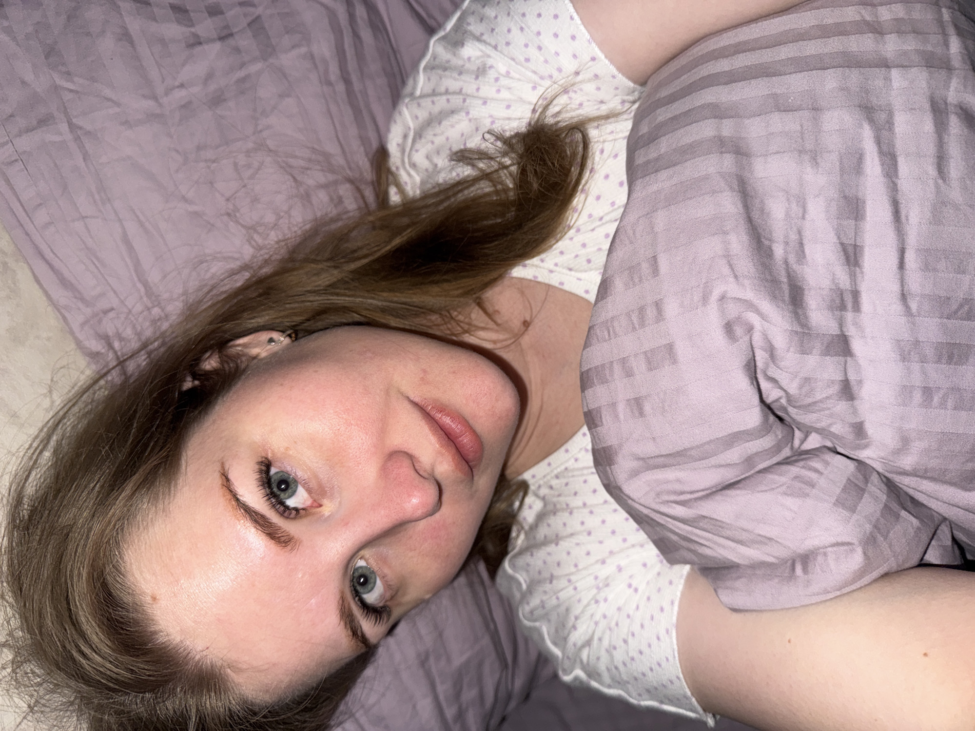 A woman with fair skin, light brown hair, and blue eyes is lying in bed. She is partially covered by a light purple blanket with horizontal stripes. She is wearing a white shirt with light purple polka dots. The bedsheets are a similar light purple color. The location is unknown but specified as possibly being in Gornyi (Dvurechenskoe S/P), Russia, though there is no way to confirm this from the image.