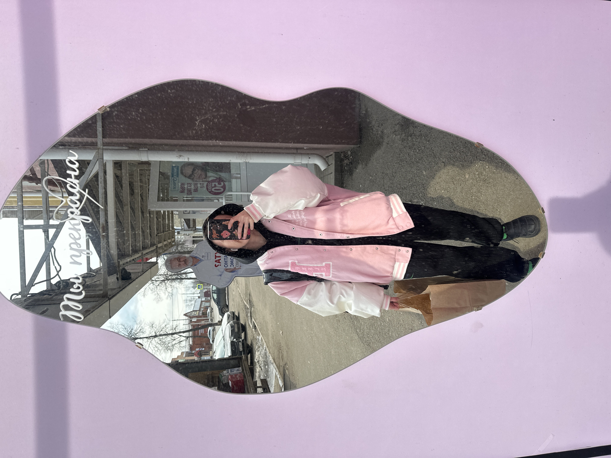 The image shows a young person taking a mirror selfie in a public location in Votkinsk, Russia. The mirror, shaped like lips, is attached to a pale lavender wall, reflecting a street scene. The person is wearing a pink and white varsity jacket, black pants, and black shoes with green accents. They are holding a smartphone to capture the selfie. Reflected in the mirror is a street view of what appears to be a storefront with a large advertisement. In the background, cars and buildings are visible, indicating a city environment. The mirror also reflects the image of an older man standing behind the photographer in the street. He wears a white shirt with lettering that appears to advertise a political candidate. A brown paper bag is visible in the lower right corner of the mirror's reflection.