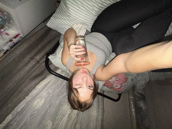 A woman lying on her back in bed takes a selfie. She wears a gray tank top, black leggings, and drinks from a clear glass with a straw. The bed has striped pillows and a floral pillow behind her back. A metal frame surrounds the bed, and a sheer, lace-patterned fabric hangs from it. To the left, a shelf contains assorted items. The image has a slightly tilted perspective.