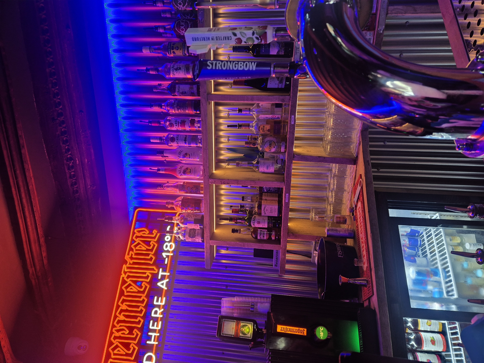This is an indoor shot of a bar area in West Suffolk, United Kingdom. The focal point is a well-stocked back bar, illuminated with vibrant red and blue neon lights. A neon sign for "Jägermeister" is prominent, indicating that the drink is served chilled at -18°C. Shelves behind the bar display an assortment of liquor bottles including Malibu, Bacardi, Captain Morgan, Strongbow Cider, Jose Cuervo, Cazcabel and Jack Daniel's. A metal dispenser for Jägermeister is visible, with a temperature display confirming it is chilled to -18°C. A refrigerator filled with canned and bottled drinks, including Red Bull and fruit juices, is also visible. Other bar elements include a Fishery ice bucket, a metal beer tap, and a metal, corrugated wall covering. There are no people visible in the image.