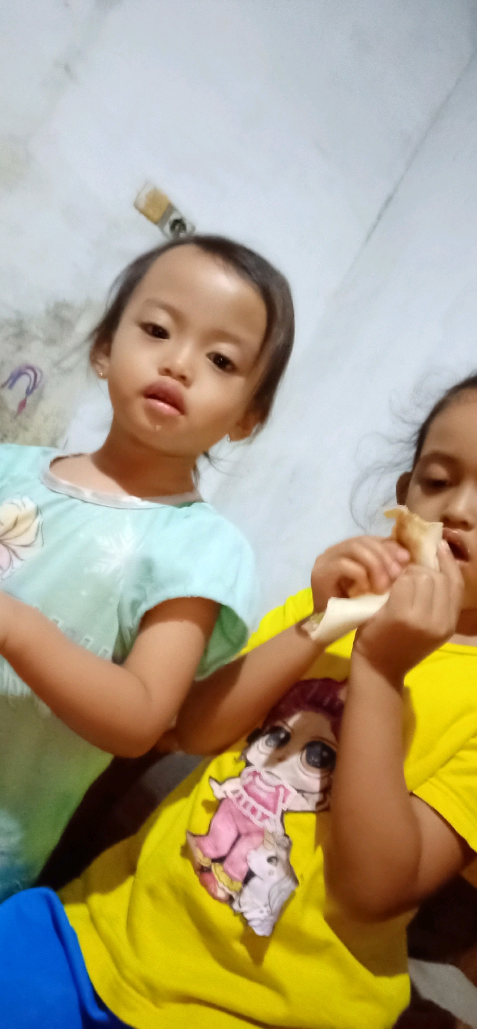 This is a close-up, eye-level, slightly angled, portrait-oriented photograph of two young girls in what appears to be an indoor setting. The background is a plain, textured wall, with some light and shadow, suggesting a dimly lit room.
The girl on the left is facing the camera directly, with a slight pout and a smudge of food on her chin. She has dark hair pulled back and is wearing a light blue t-shirt with a design featuring snowflakes and a floral pattern, possibly from the movie "Frozen." Her expression is somewhat solemn.
The girl on the right is partially visible, turned slightly away from the camera, and is in the process of eating something wrapped in a light-colored paper, possibly a snack or a piece of food. She is wearing a bright yellow t-shirt adorned with a large graphic of a doll's face with big eyes, a common style of popular toy dolls. Her focus is on the food she is eating.
The lighting appears to be artificial, casting soft shadows. The overall mood is casual and domestic, capturing a candid moment of the girls sharing a snack. There is no visible text in the image. The location context provided is Bojanegara, Indonesia.