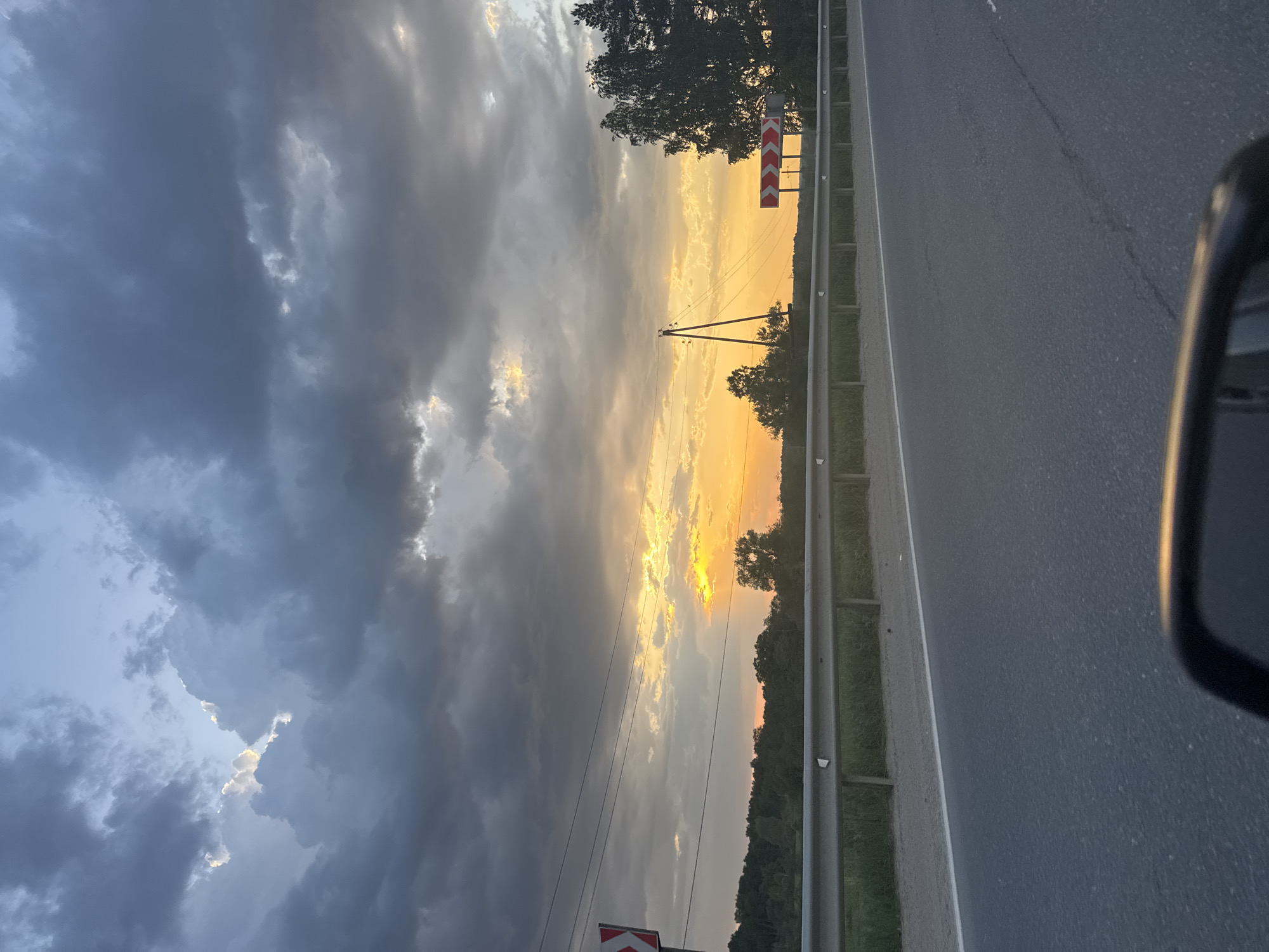 The image depicts a scene near Раздолье, Russia, at dusk or dawn. A dark asphalt road occupies the right side of the frame, bordered by a metal guardrail. Beyond the guardrail is a strip of vegetation followed by a denser tree line along the horizon. In the sky, heavy grey clouds dominate the left side of the image. A bright yellow/orange glow pierces through the clouds and illuminates the sky above the tree line. A utility pole with wires is visible extending into the sky. A red and white chevron sign is on the left side of the guardrail. A portion of a car mirror is visible in the lower right corner of the frame.