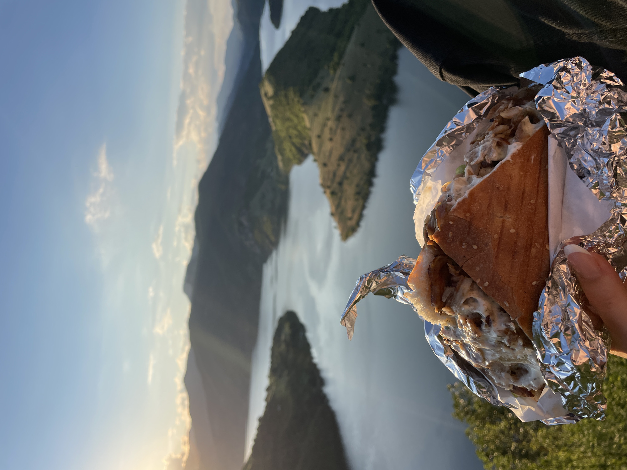 In Borovitsa, Bulgaria, a person holds a sandwich wrapped in aluminum foil in the foreground. The sandwich appears to be a large, triangular piece of bread filled with meat and a creamy sauce, with sesame seeds on the crust. In the background, a vast, winding body of water, likely a dam or a lake, is visible, surrounded by rolling, tree-covered hills and mountains. The sun is setting, casting warm rays of light across the landscape and creating a serene, natural scene.