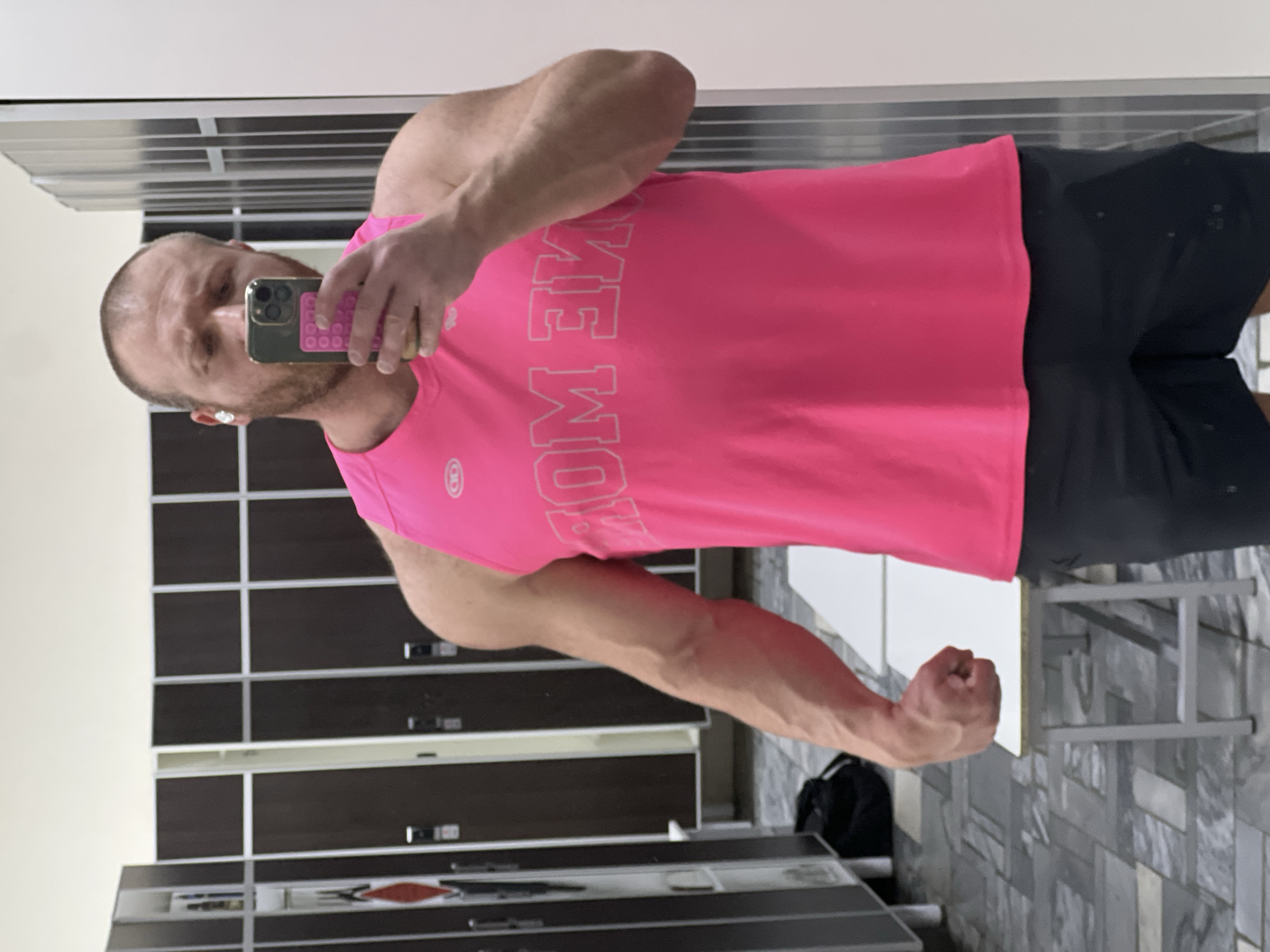 This image depicts a male individual taking a mirror selfie in a locker room, likely within a fitness or sports facility in Chaikovsky, Russia. The man has a muscular build, short light-brown hair, and a beard. He is wearing a bright pink sleeveless athletic top with white, mirrored text across the chest (appearing as "MOM MEW"), black athletic shorts, and a white wireless earbud in his right ear. He holds a gold-colored smartphone, equipped with a bright pink 'pop-it' style case, in his right hand, angled to capture his reflection. His left arm is flexed, displaying prominent veins, and his left hand is clenched into a fist, resting on a white, narrow bench with light-colored metal legs. The background features rows of dark brown and light grey metal lockers, some with horizontal ventilation slats, lining the walls. The floor is covered in light and dark grey square tiles arranged in a patterned manner. A black duffel bag is visible on the floor near the lockers on the left side of the frame.