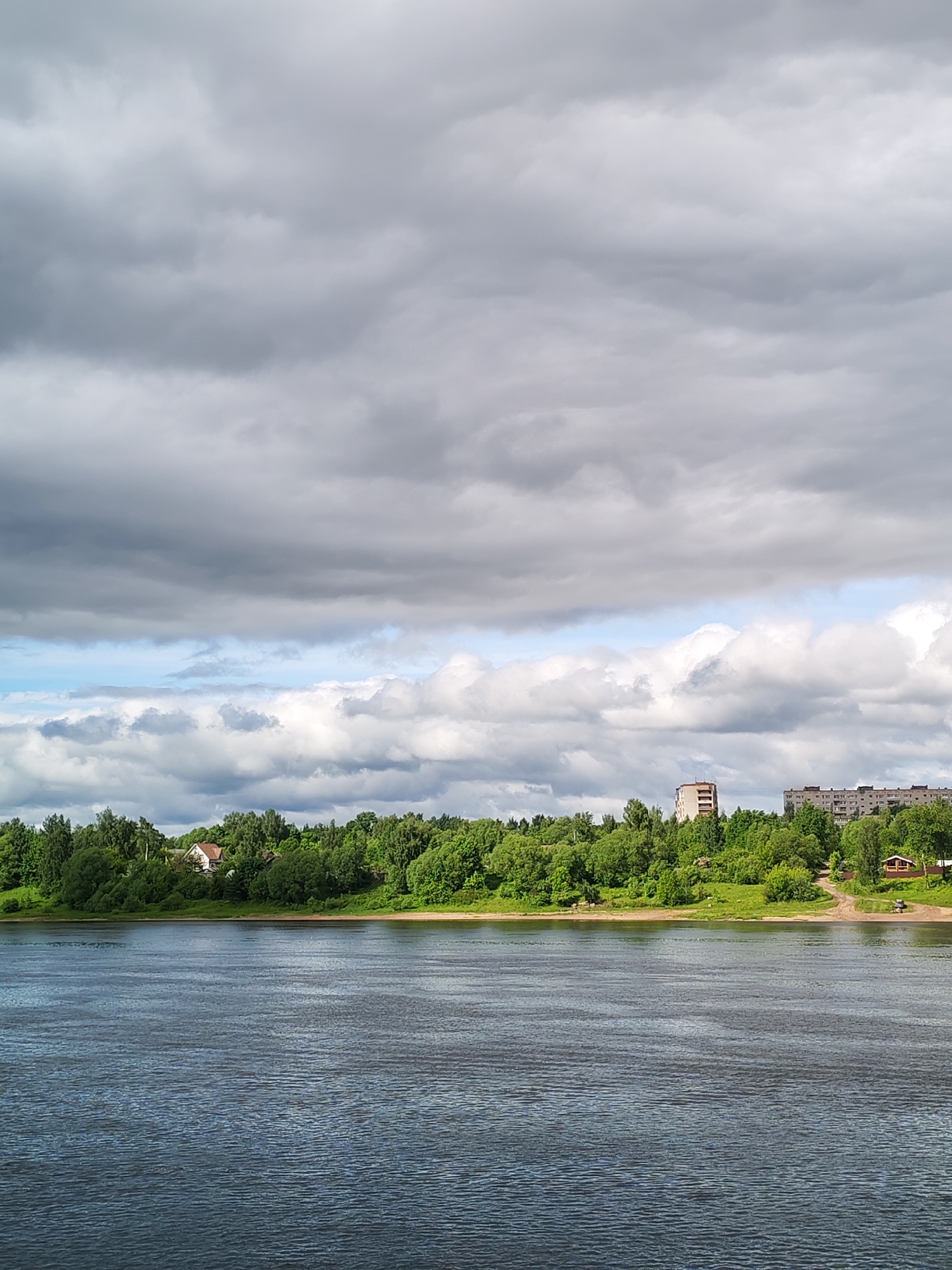 The image depicts a body of water, likely the Volga River, with a treeline and buildings visible on the opposite shore. The sky is overcast with heavy clouds. The buildings on the shore include apartment blocks and a single, taller structure that could be a hotel or office building. A single, small house is visible amid the trees on the left. The location is likely Rybinsk, Russia, given the style of the buildings and the presence of the Volga River. There is no visible human activity in the image.