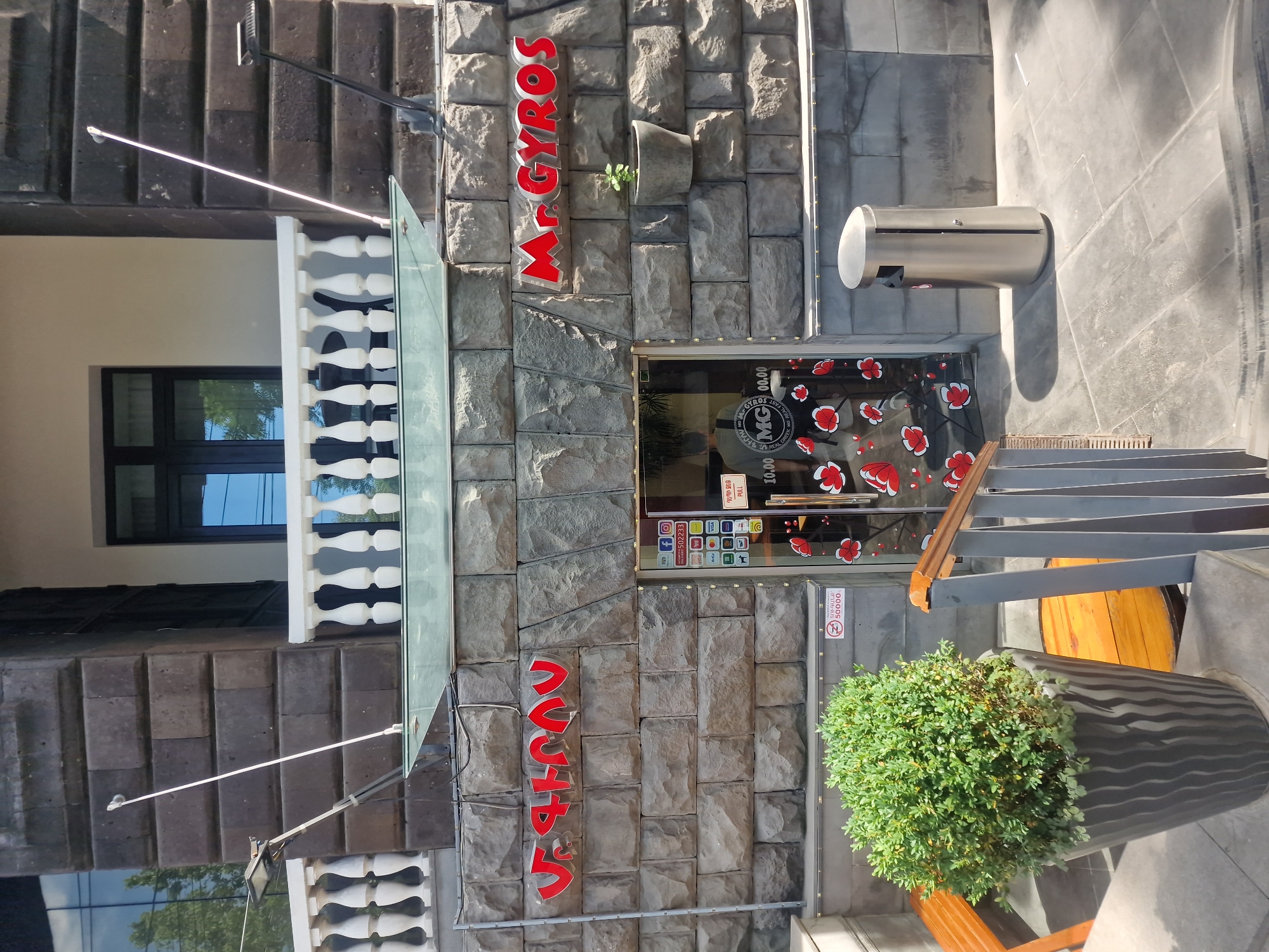 This media file shows the exterior of a restaurant called "Mr. Gyros" in Yerevan, Armenia. The building facade is made of grey stone blocks. Red illuminated letters spell out "Mr. GYROS" vertically on the left side of the entrance, and "ՏՈՒԳԱՆՔ" (likely a local language word) is written vertically in red on the right. The entrance features a glass door decorated with red floral patterns, and a glass awning above it.
A stainless steel trash receptacle is mounted on the wall to the right of the entrance. Social media icons and payment method logos are visible on a sign on the glass door. A potted green plant is situated in front of the building, next to a staircase. The scene appears to be set during the day, under sunny weather conditions, with shadows indicating sunlight. The overall impression is of a casual dining establishment.