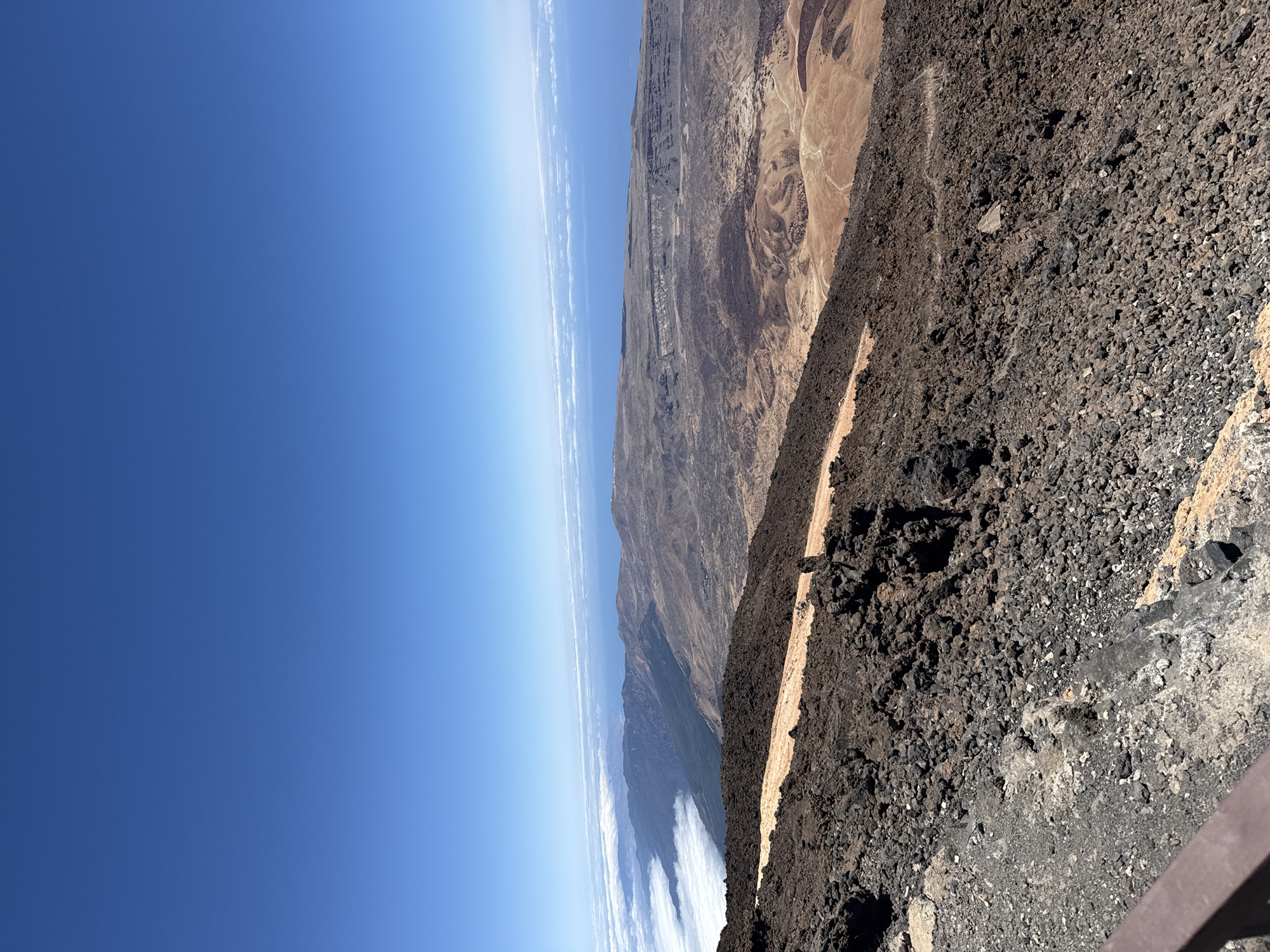 The image depicts a panoramic view of a volcanic landscape with a vast expanse of blue sky. In the foreground, dark, rocky volcanic terrain dominates, with a narrow, lighter-colored path winding through it. In the mid-ground, the landscape transitions to a more arid, reddish-brown terrain with undulating hills and valleys, exhibiting various shades of brown and orange. A significant geological feature, possibly a crater rim or caldera wall, is visible in the distance, characterized by layered rock formations. Further into the distance, a layer of white clouds stretches horizontally, resembling a sea of cotton, and beyond that, the deep blue of the ocean meets the horizon. The clear, cloudless sky above suggests a sunny day. The scene appears to be a high-altitude vantage point, likely within the Teide National Park, which is located in the municipality of La Orotava on the island of Tenerife, Spain. There are no people or animals visible in the image.