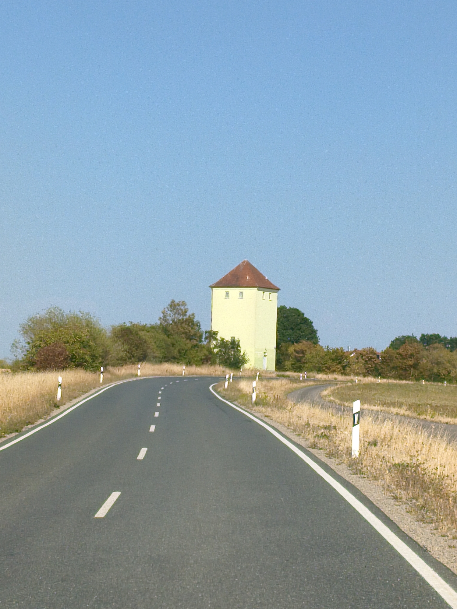 A light yellow, square tower with a red-tiled, gabled roof stands prominently on a slight rise to the right of a curving asphalt road. The tower has several small, evenly spaced windows on its visible facade and appears to be a historic building, possibly a water tower or observation tower. The road itself is two-lane with a dashed white line marking the center and a solid white line along the right edge, suggesting it is a rural or suburban thoroughfare. White and black striped road markers are visible along the sides of the road. The landscape surrounding the road is characterized by dry, golden grass and scattered trees with autumn foliage. The sky is a clear, bright blue with no visible clouds. The scene captures a serene, sunlit day in the German countryside, specifically in or near Markt Erlbach, Germany, given the tower's architectural style and the surrounding environment, which are consistent with the region. There are no people or animals visible in the image, and no actions or interactions are depicted.