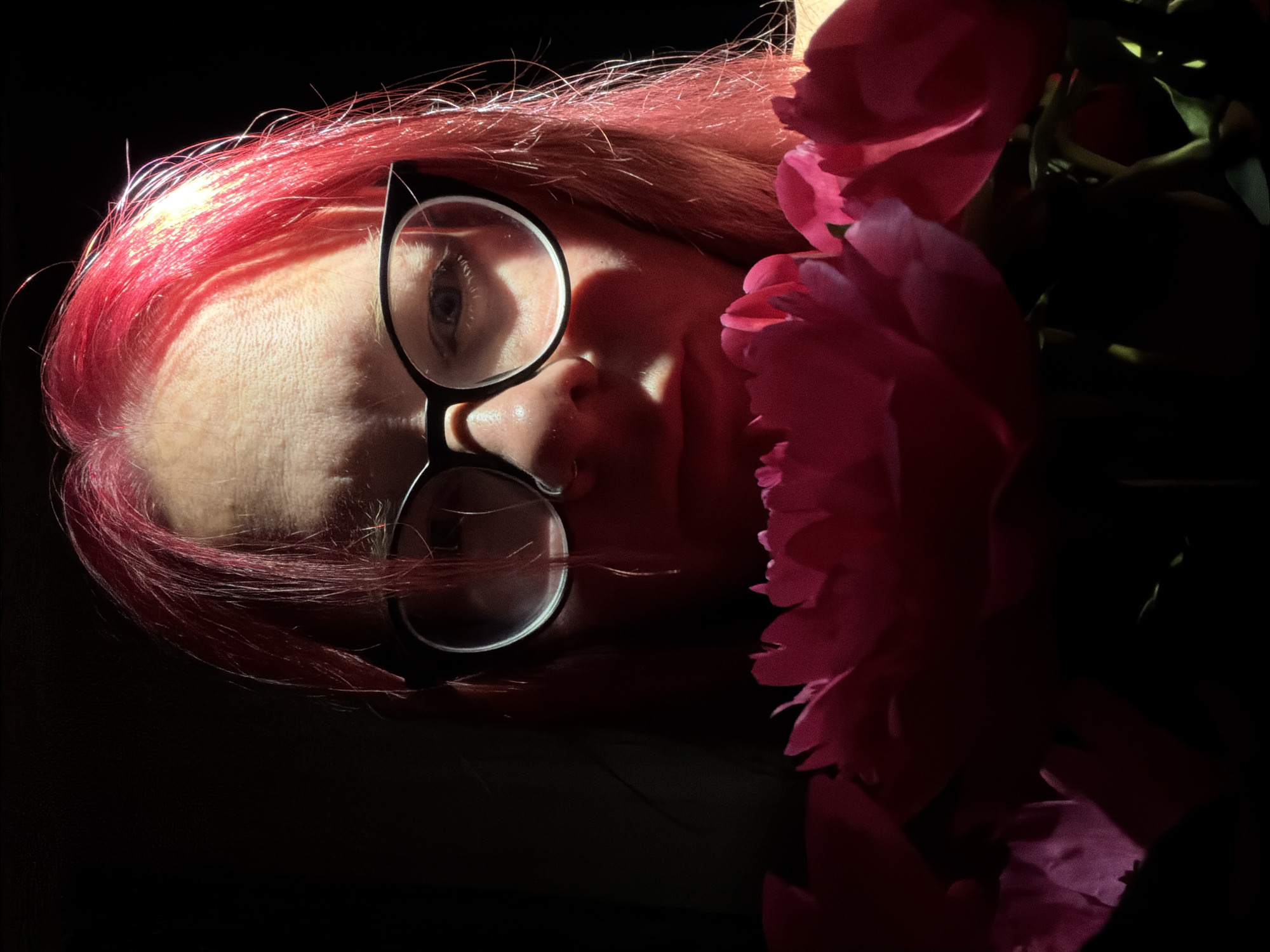 A person with vibrant, bright pink hair is depicted in a high-contrast, close-up shot against an entirely dark, featureless background. Their face is partially illuminated by a strong light source positioned above and to the viewer's left, creating significant highlights on the forehead, the right side of the face, and the hair. Deep shadows obscure the left side of their face and the area beneath their nose and chin. The individual wears black-framed glasses with clear lenses; their light-colored eyes are visible through the lenses, directed straight forward. A cluster of large, deep pink or magenta flowers, likely peonies, occupies the lower foreground, partially obscuring the person's mouth and the lower portion of their face. The petals of the flowers are layered and soft, catching some of the overhead light. No specific location, including the city of Chaikovsky, Russia, is discernible from the image content.