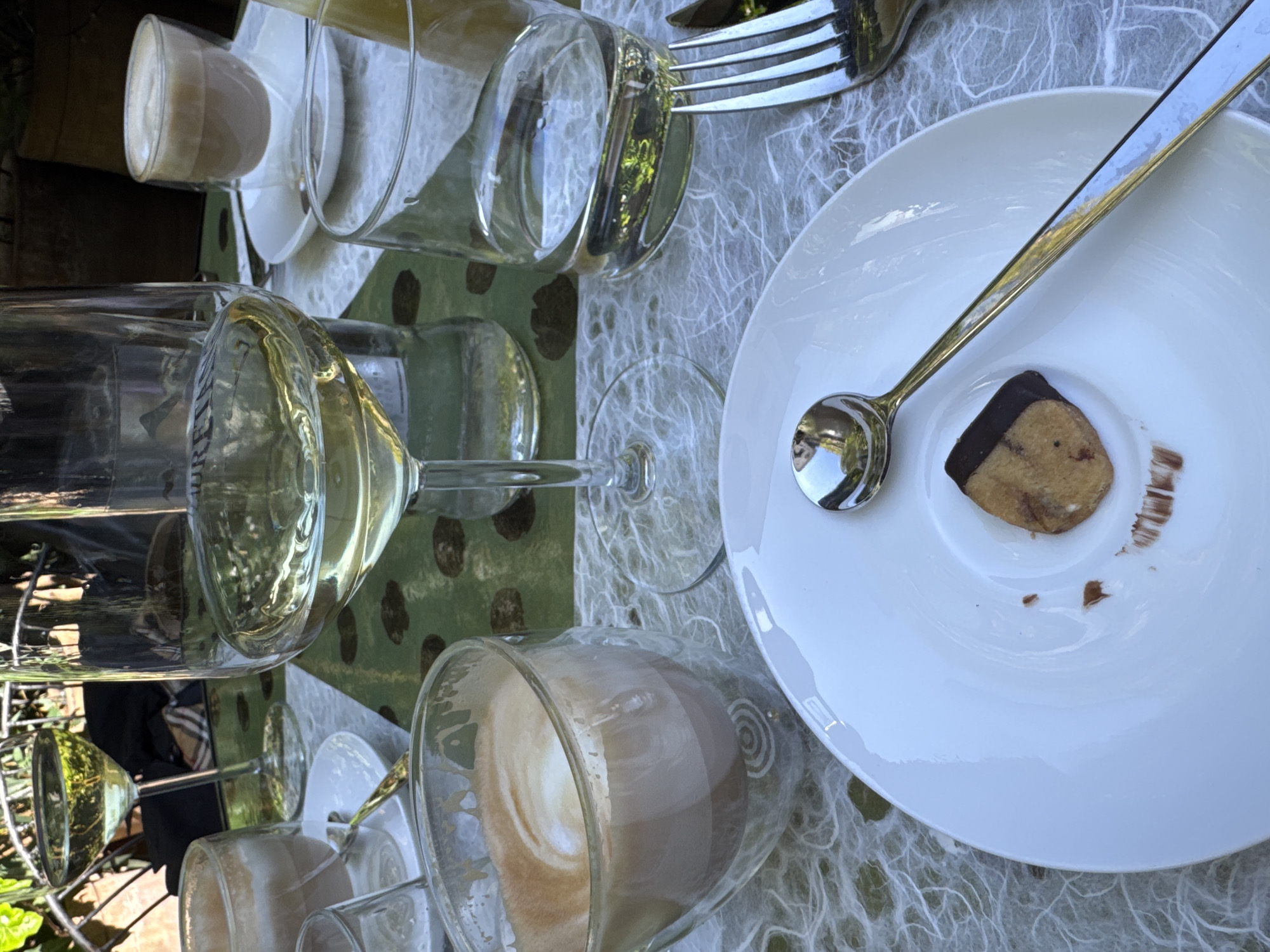 This media file captures an outdoor table setting, viewed from a slightly elevated perspective. The scene is bathed in bright, natural daylight, suggesting it's either morning or afternoon. The table is adorned with a layered arrangement of textiles: a green mat with dark brown, irregular patterns, topped by a translucent, white, fibrous placemat or tablecloth.
The table is set with multiple clear glass beverages and a dessert. There are at least two wine glasses, one holding a clear liquid possibly white wine, and another empty. Several other glasses contain milky coffee drinks, likely lattes or cappuccinos, with distinct latte art visible in one. Another tall glass holds a lighter-colored, possibly yellowish, drink.
A pristine white ceramic plate sits prominently on the right side, holding a single, small, light-brown cookie partially covered in dark chocolate. A silver spoon rests diagonally across the plate. A silver fork is also visible towards the top right, partially obscured by a glass.
In the background, lush green foliage is discernible, indicating an outdoor or garden setting. A person is faintly visible in the mid-background, wearing an item of clothing with a distinct brown and beige plaid pattern. The overall atmosphere appears relaxed and leisurely, suggesting a casual gathering, brunch, or coffee break, possibly at a cafe or restaurant patio in Milan, Italy. Faint, illegible text can be seen on the side of one of the clear glasses.