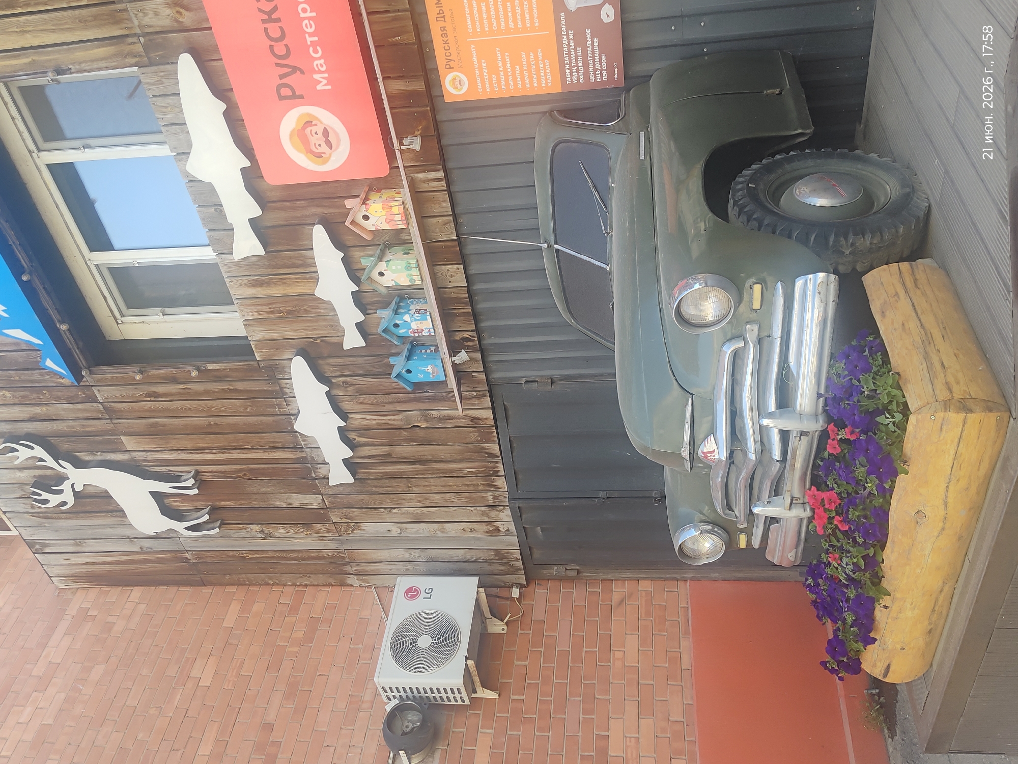 The image shows a vintage green car parked in front of a building with a wooden facade. The car has chrome detailing on the grille and headlights, and it is positioned in front of a wooden planter filled with purple and pink flowers. The building's facade features a silhouette of a deer and several stylized white fish cutouts. Above the car, a window is visible. A sign with Russian text and a drawing of a man with a mustache is displayed on the right side of the building. An air conditioning unit is mounted on the brick wall to the left of the wooden facade. The timestamp "21 июня. 2026 г., 17:58" is visible in the bottom right corner of the image, suggesting the photo was taken in Astana, Kazakhstan.