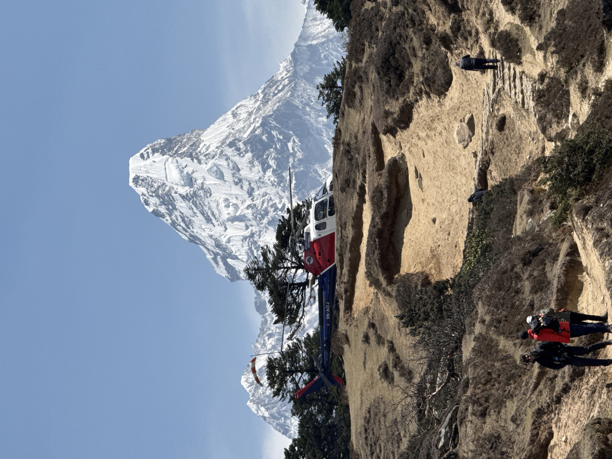 The scene, likely in Khumjung, Nepal, shows a red and blue helicopter parked near the base of a snow-covered mountain. A rocky cliff face occupies the right side of the image, with sparse vegetation growing on it. Several people are walking along a narrow path carved into the cliff. The sky is clear and blue.