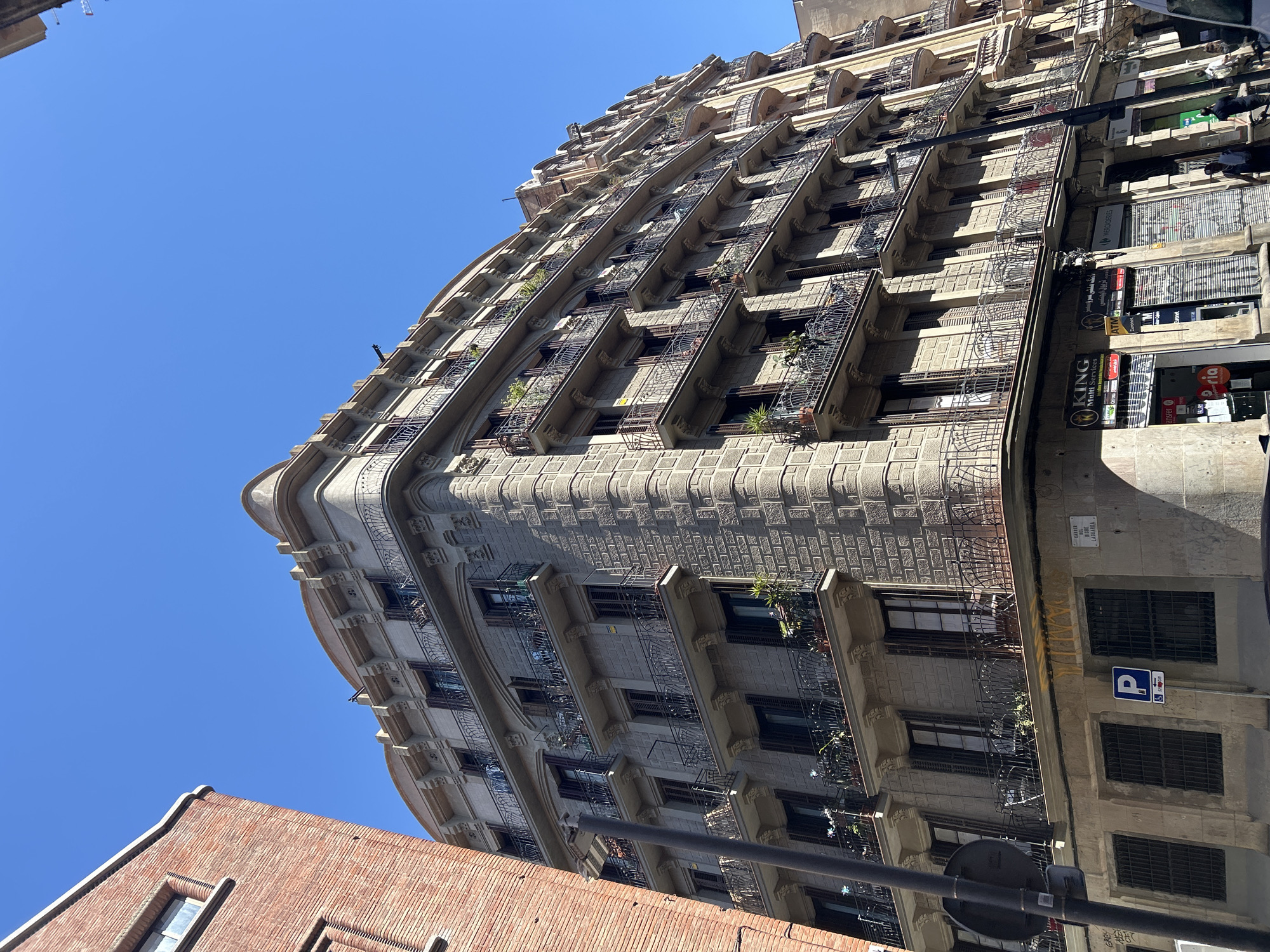 This media file captures an upward view of a tall, ornate building in an urban setting under a clear blue sky. The building, likely of historic architectural style, features a distinctive curved corner and numerous balconies with intricate wrought-iron railings. Its stone facade is textured with a regular, block-like pattern, and decorative elements are visible below the balconies and along the roofline.
Many balconies host potted plants, adding greenery to the grey stone. Windows are generally shuttered or have dark panes. The bright sunlight casts distinct shadows, indicating a sunny day, likely mid-morning or early afternoon.
At street level, the building accommodates commercial establishments. Several signs are visible, including "KING Multi Services," "ATM," "transfer," and an Arabic script sign reading "تريول ايجنسي" (Travel Agency). Another sign displays "MERCADERIES." A European-style blue "P" parking sign is also present, alongside a street sign that clearly reads "CARRER DEL BISBE LADUARDA," confirming the location as Barcelona, Spain. A darker, plainer brick building is partially visible in the foreground, to the left, contrasting with the ornate main structure. A black street pole and partial street lamp obscure some of the lower details.