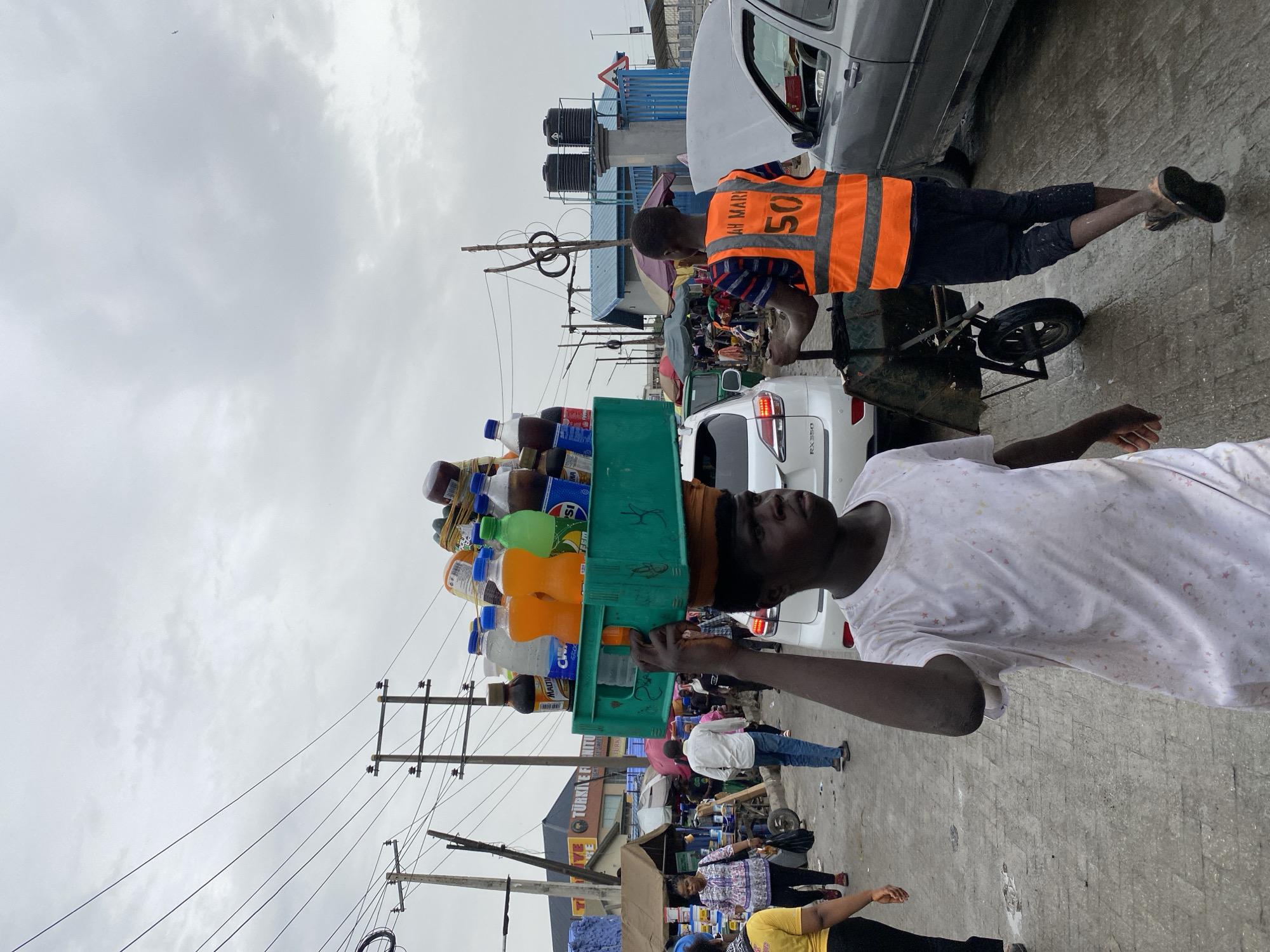 The image shows two men in Lagos, Nigeria, engaged in commerce on a street. One man carries a large stack of drinks and containers on his head, supported by a green plastic crate. He wears a white t-shirt with a star pattern. The second man pushes a cart with assorted items. He wears an orange vest labeled "AH MARI 50". They are both walking on a paved street. Cars are parked nearby, including a white RX350. Overhead utility lines are visible against an overcast sky. In the background, there is a sign that reads "TURKIYE FI INITU". Other people walk along the street in the background, near a commercial area.