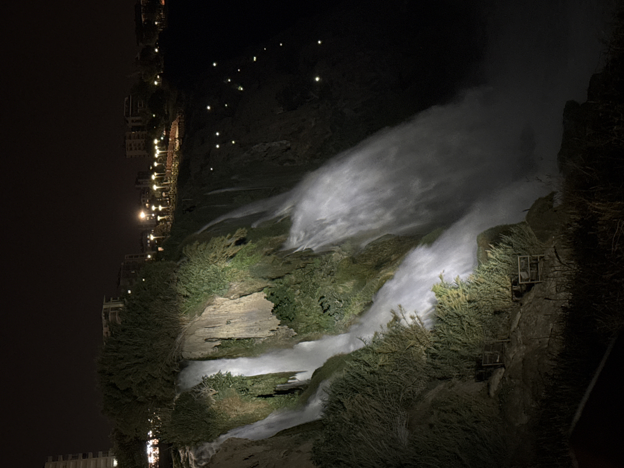 This is a nighttime, high-angle, vertical shot of the Duden Waterfalls in Antalya, Turkey. The waterfall cascades down a rocky cliff face covered in lush green vegetation. The water is illuminated by artificial lights, creating a glowing effect as it falls. In the background, there are several buildings and a park area with string lights. The overall impression is one of natural beauty enhanced by urban elements.