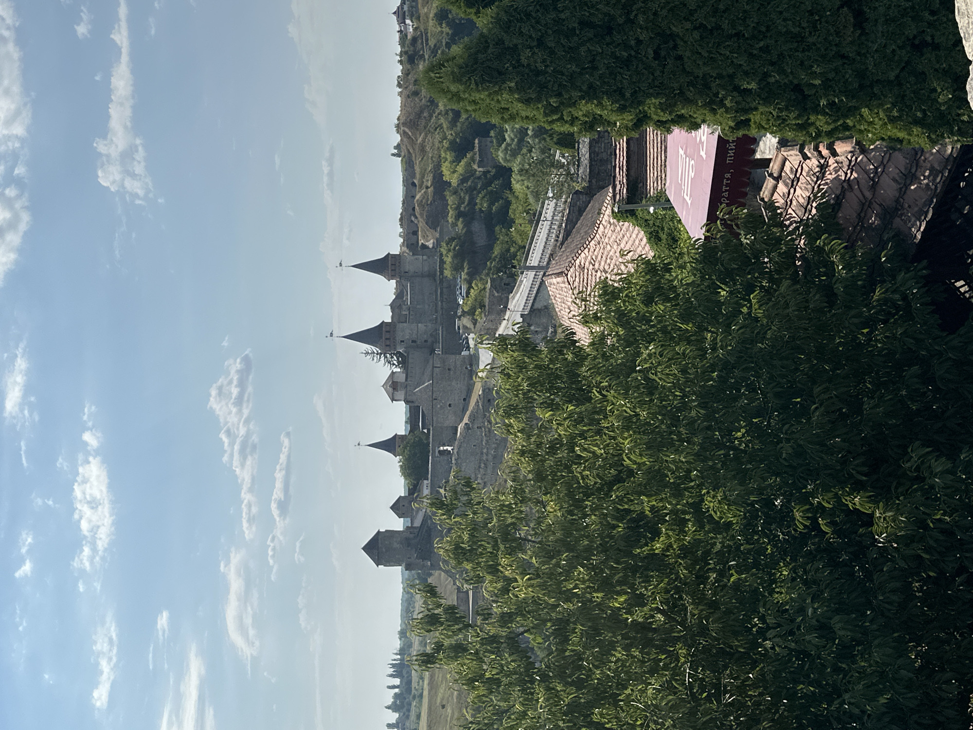 This image captures a view of the Kamianets-Podilskyi Castle in Kamianets-Podilskyi, Ukraine, on a clear day with scattered clouds. The castle, a prominent historical fortress, is situated on a rocky promontory, its stone towers and ramparts visible against the sky. In the foreground, lush green trees partially obscure the view, with a section of a red-tiled roof and a maroon awning with white lettering visible in the lower right portion of the frame. A bridge can be seen extending towards the castle complex, and the surrounding landscape appears hilly and vegetated. There are no people clearly visible in the image.