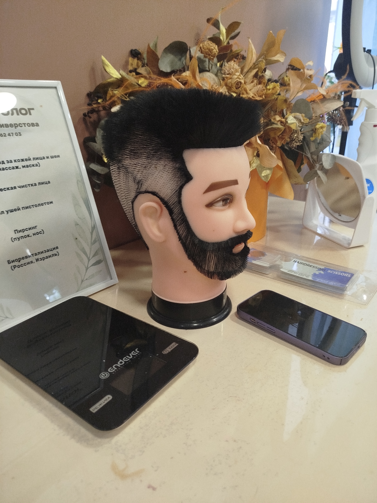 The image shows a mannequin head with a stylized haircut and beard, positioned on a table. To the left of the mannequin, there is a framed certificate or document with text in Russian. In front of the mannequin, there is a pair of hairdressing scissors in a clear plastic case. A smartphone is placed on the table to the right of the scissors. A digital scale is visible in the lower left corner of the image. In the background, a bouquet of dried flowers and a spray bottle are present. The text on the document appears to be related to cosmetic procedures, mentioning "уход за кожей лица и шеи" (face and neck skin care), "массаж, маска" (massage, mask), "мезотерапия" (mesotherapy), and "биоревитализация (Россия, Израиль)" (biorevitalization (Russia, Israel)). This suggests the setting is likely a beauty salon or a similar establishment. No specific landmarks or street views are visible to pinpoint the exact location in Pskov, Russia, beyond the text on the document indicating services offered which originate from Russia and Israel.