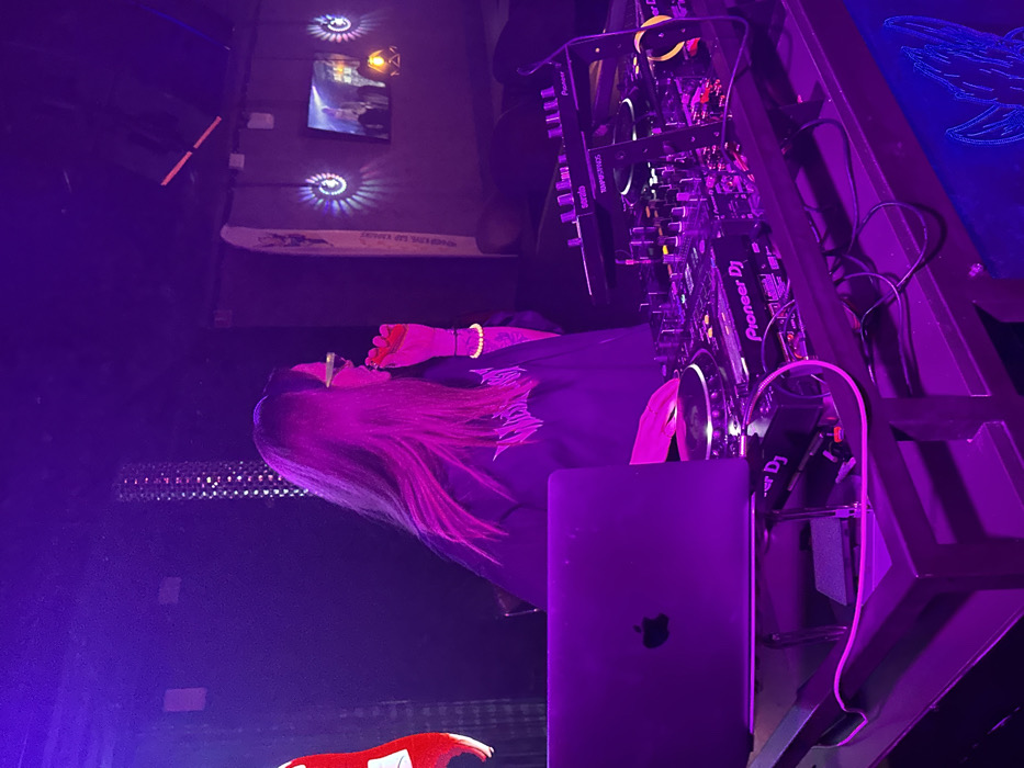 A DJ stands next to their equipment, including a Pioneer DJ setup and a MacBook. The scene is lit with purple hues, suggesting a club or event setting. The DJ has long hair and is holding something near their face. Above, there are light fixtures and a framed image on the wall. The focus is on the DJ and the equipment, highlighting a performance or event.