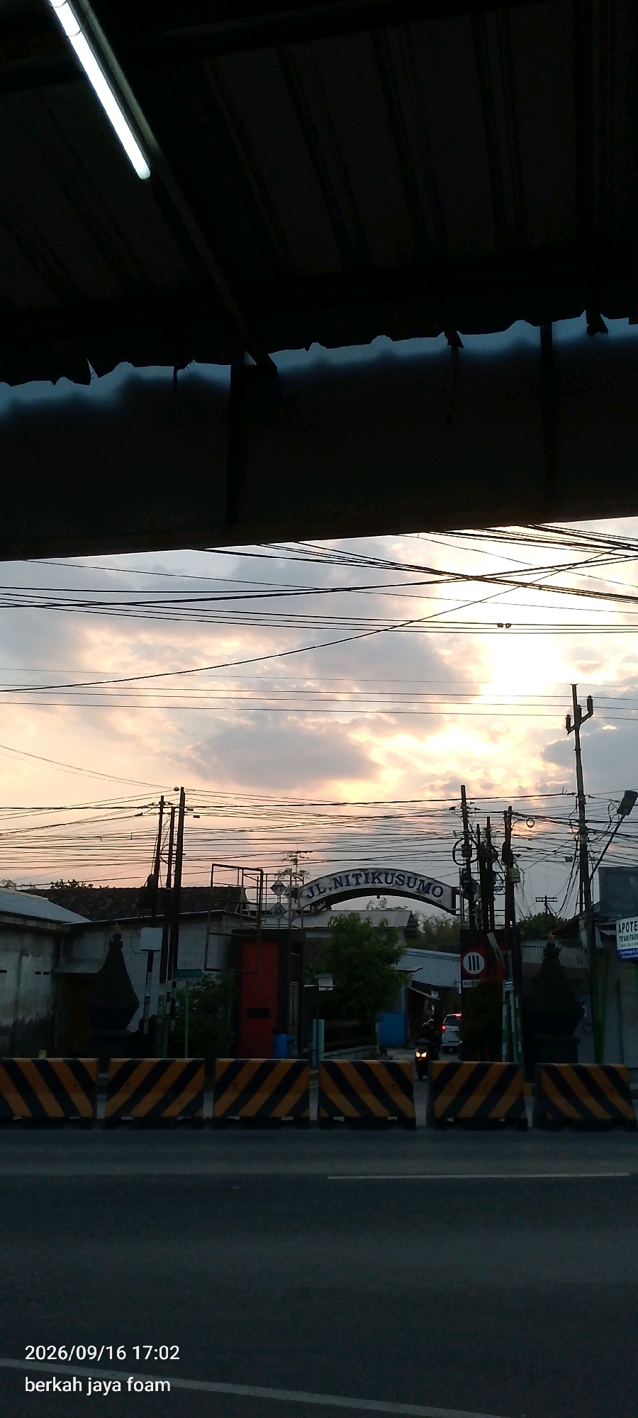 The image captures an outdoor scene in Madiun, Indonesia, likely at dusk. In the foreground, a paved road is visible, bordered by a row of black and yellow striped traffic barriers. Above the road, a complex network of overhead power lines stretches across the frame, silhouetted against a cloudy sky illuminated by the warm hues of sunset.
The middle ground reveals a street with buildings on either side. A prominent arched gateway with the text "JL. NITIKUSUMO" is centered, suggesting this is a street or entrance. A traffic sign with the Roman numeral "III" in a circle is also visible near the gateway. Several utility poles with numerous wires are scattered throughout this area. A few vehicles, including a motorcycle with its headlight illuminated and a car, are present on the road. On the left side of the street, a building with a dark, possibly red, door is noticeable. A few people, appearing as silhouettes, are seen standing or moving in the vicinity of the buildings.
The background shows more buildings and trees, fading into the atmospheric lighting of the sunset. The upper portion of the image is dominated by a dark, shadowed structure, possibly an overhang or roof, with a fluorescent light fixture attached to it, casting a glow. The timestamp "2026/09/16 17:02" and the text "berkah jaya foam" are superimposed on the lower left corner of the image, indicating a date and a potential business name.