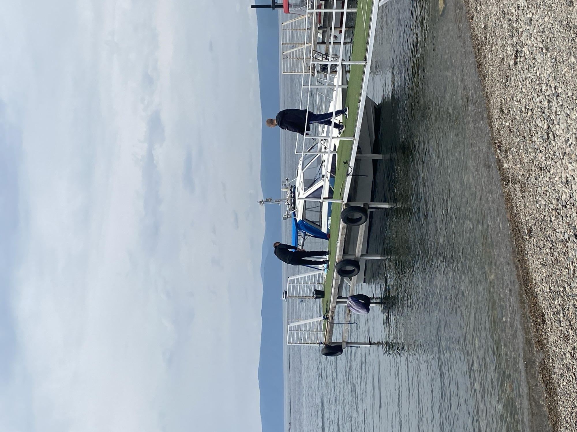 Two men are on a pier next to a boat in Listvyanka, Russia. One man in a black jacket and dark pants is standing on the pier and appears to be cleaning a piece of equipment on the boat. The other man, wearing a black jacket and blue jeans, is walking away from the boat on the pier. The boat is white with a blue canopy and the word "YAMAHA" is visible on its side. The pier is made of metal with white railings and artificial green turf. There are several black tires attached to the pier, serving as fenders. The water is calm and reflects the sky and the pier. In the background, there are mountains and a cloudy sky. The foreground shows a pebbly shoreline.