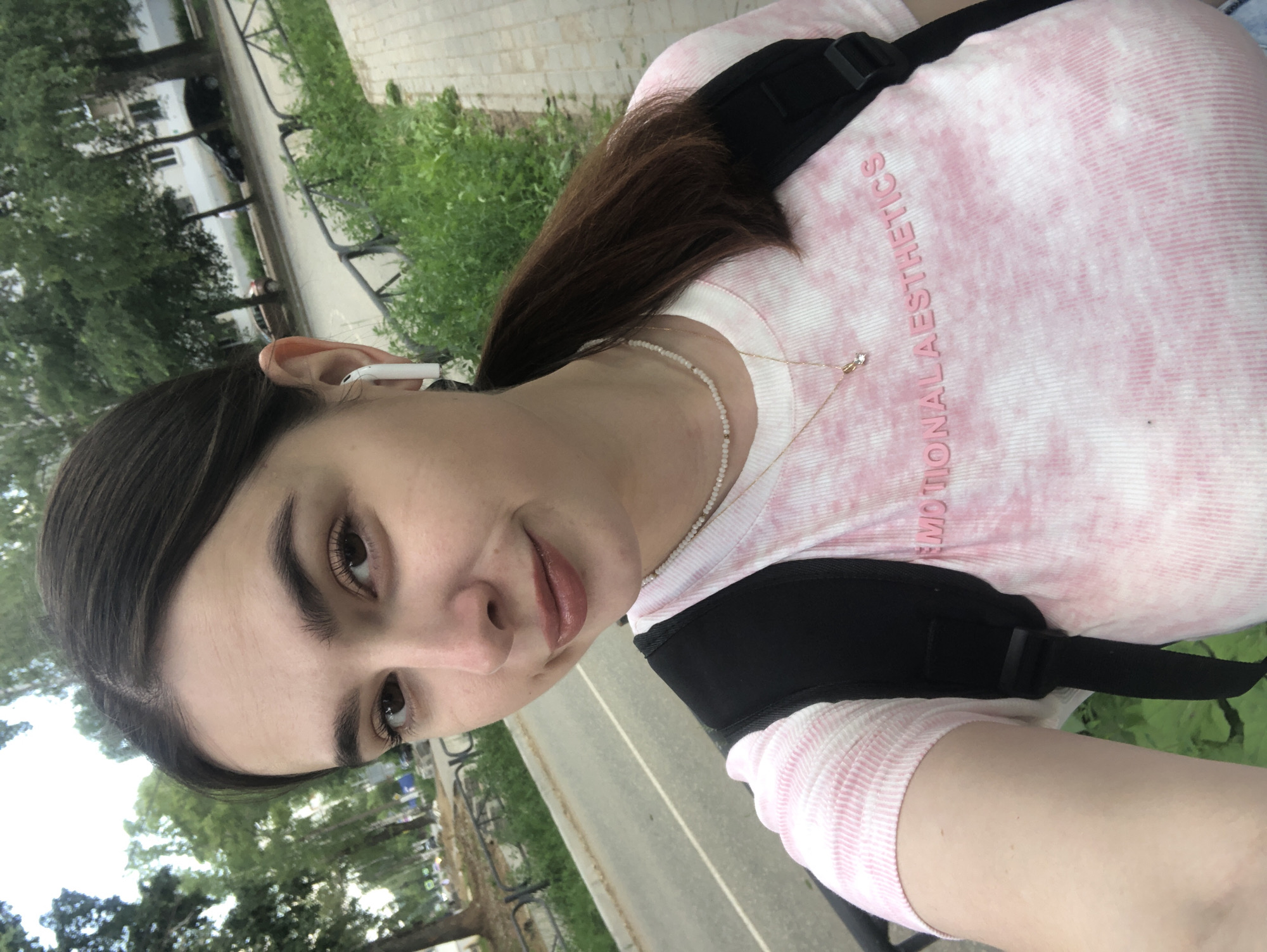 The image shows a young woman with dark hair and brown eyes taking a selfie outdoors. She is wearing a pink and white tie-dye t-shirt with the text "EMOTIONAL AESTHETICS" printed on it, a thin pearl necklace, and a small gold necklace with a pendant. She has white earbuds in her ears and is wearing a black backpack. The background suggests an urban setting with trees, a sidewalk, a road with cars, and buildings visible in the distance. The overall impression is of a casual, everyday scene, likely on a street or path in the city of Chaikovsky, Russia.