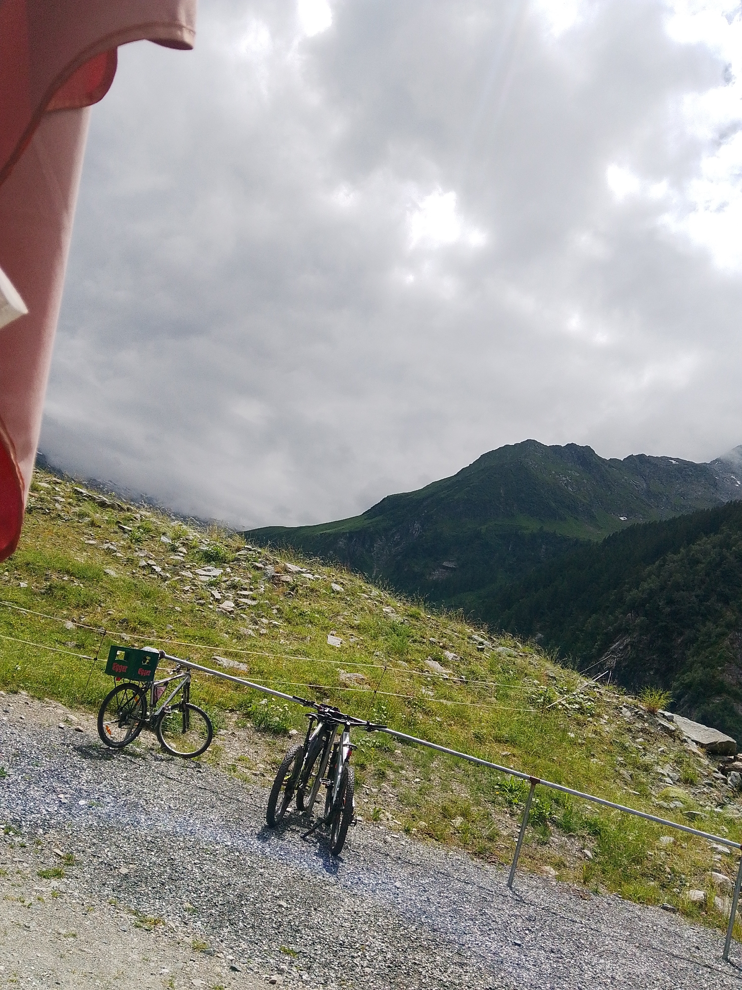 The image shows a scenic mountain landscape in Bramberg am Wildkogel, Austria. In the foreground, two bicycles are parked on a gravel path. One bicycle, positioned further left, is a silver mountain bike with a green beer crate strapped to its rear rack. The crate displays the text "Egger" and "Gleisdorf" in white lettering. The second bicycle, to the right, is a dark-colored mountain bike with its handlebars angled. Both bicycles appear to be parked or waiting.
The foreground path leads up a grassy, rocky slope dotted with small wildflowers. The slope is bordered by a metal railing. In the background, lush green mountains rise dramatically, with darker, forested slopes in the mid-ground. The sky is overcast with dramatic, cloudy formations, suggesting a day with mixed weather. Sunlight filters through the clouds, illuminating parts of the landscape. The overall atmosphere is serene and outdoorsy, evoking a sense of adventure and natural beauty. The time of day appears to be daytime, likely late morning or afternoon, given the lighting conditions. There are no people visible in the image, implying a moment of quiet contemplation or a pause in outdoor activity.