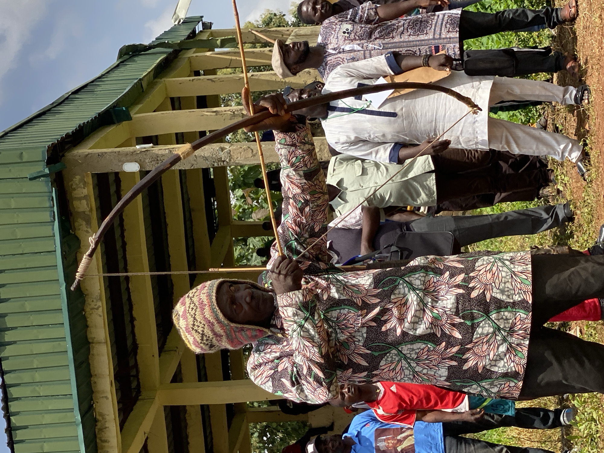 The image shows a group of people, likely in Maigizo, Nigeria, participating in an archery event or demonstration. The primary subject is an archer wearing a patterned hooded jacket and a knitted hat, drawing back a bow and arrow. Behind him stand several other individuals, some in traditional attire. The setting appears to be an outdoor area with a building featuring a green corrugated metal roof in the background. The ground is covered in grass. The composition is vertical.