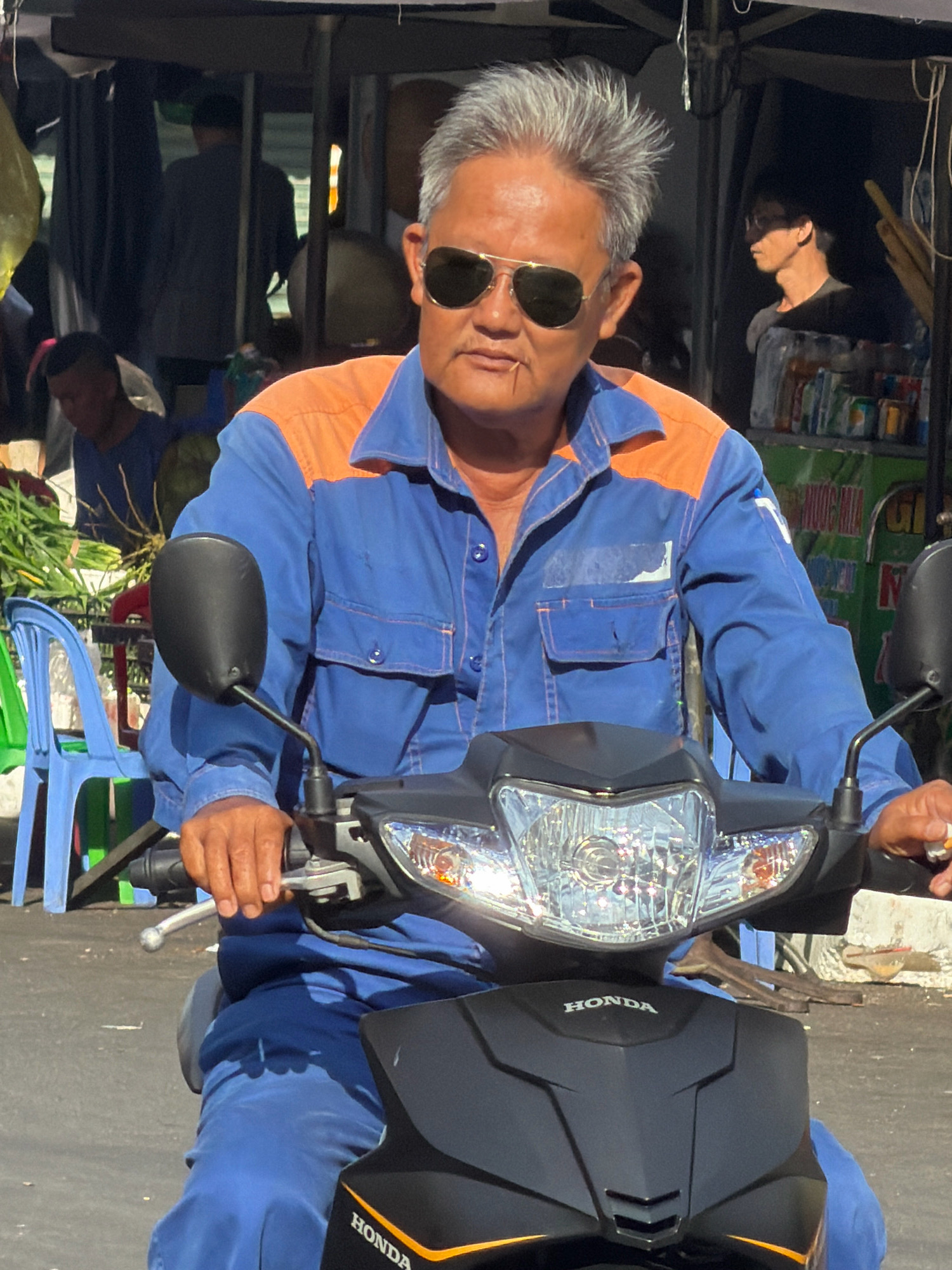 A man is riding a black Honda scooter in Nha Trang, Vietnam. He is wearing a blue work uniform with orange accents on the shoulders and collar, sunglasses, and has grey hair. He has a toothpick in his mouth. In the background, other people are visible, as well as market stalls and some green foliage. The scooter has a clear windshield and a prominent headlight. The man's hands are on the handlebars, and he appears to be looking forward. The scene is lit by bright sunlight, indicating it is daytime.